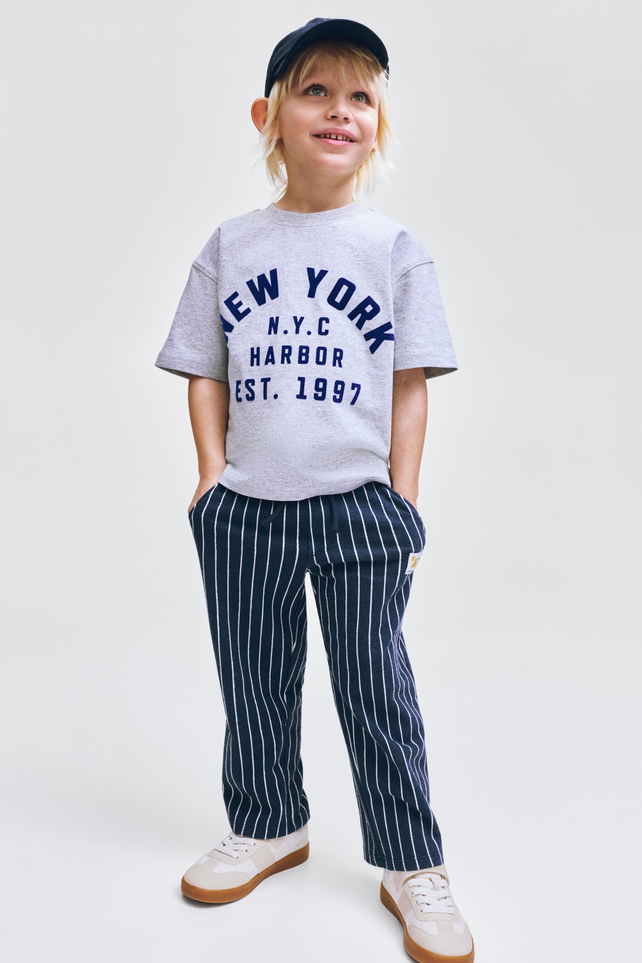 Se større billede: En grå t-shirt med korte ærmer har en rund halsudskæring og blå tekst på forsiden, hvor der står "NEW YORK N.Y.C HARBOR EST. 1997." Mørkeblå bukser viser hvide lodrette nålestriber overalt, med en lille gul lap synlig på højre hofte. En mørkeblå baseballkasket har en struktureret skygge, mens lyse sneakers viser hvide snørebånd og gummisåler.