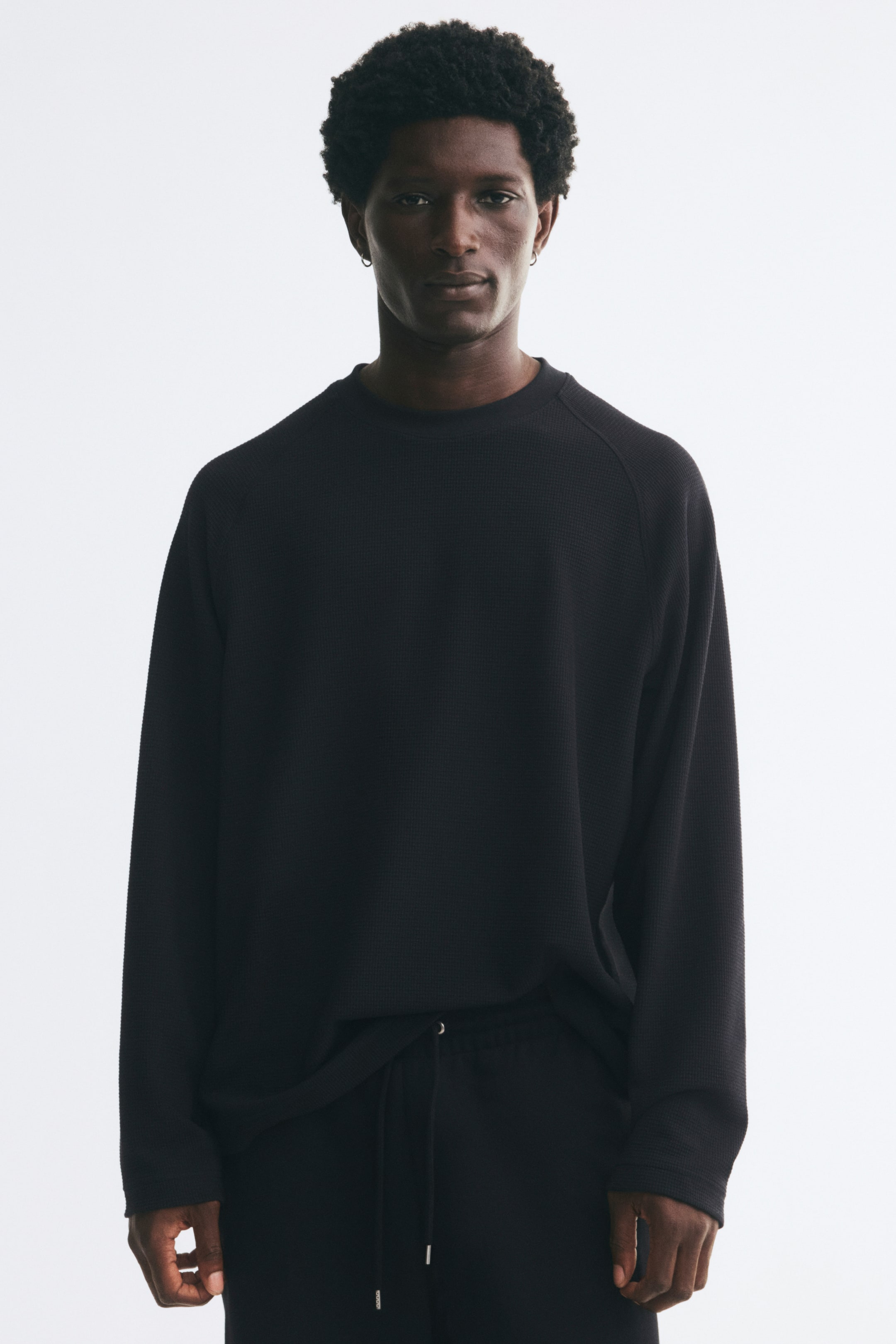 Voir une image plus grande: Un t-shirt en jersey noir texturé à motif gaufré, à coupe décontractée, avec encolure ronde et manches raglan longues, porté par le mannequin. Le t-shirt est agencé avec un pantalon noir assorti à cordon de serrage.