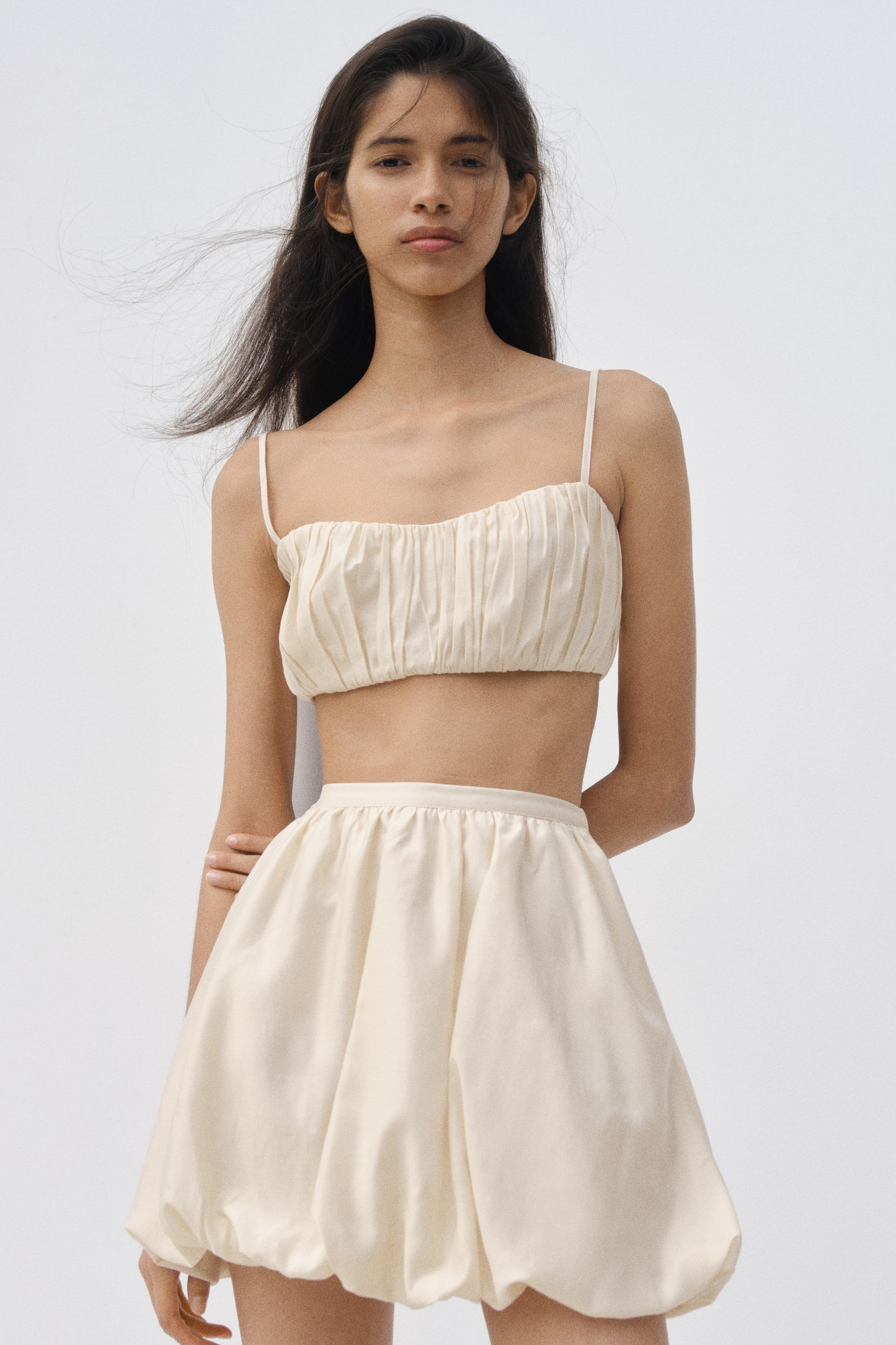 View larger image: A cream two-piece outfit features a ruched bandeau crop top with thin spaghetti straps. A matching high-waisted bubble mini skirt completes the set, showcasing a voluminous silhouette with its hem tucked under.