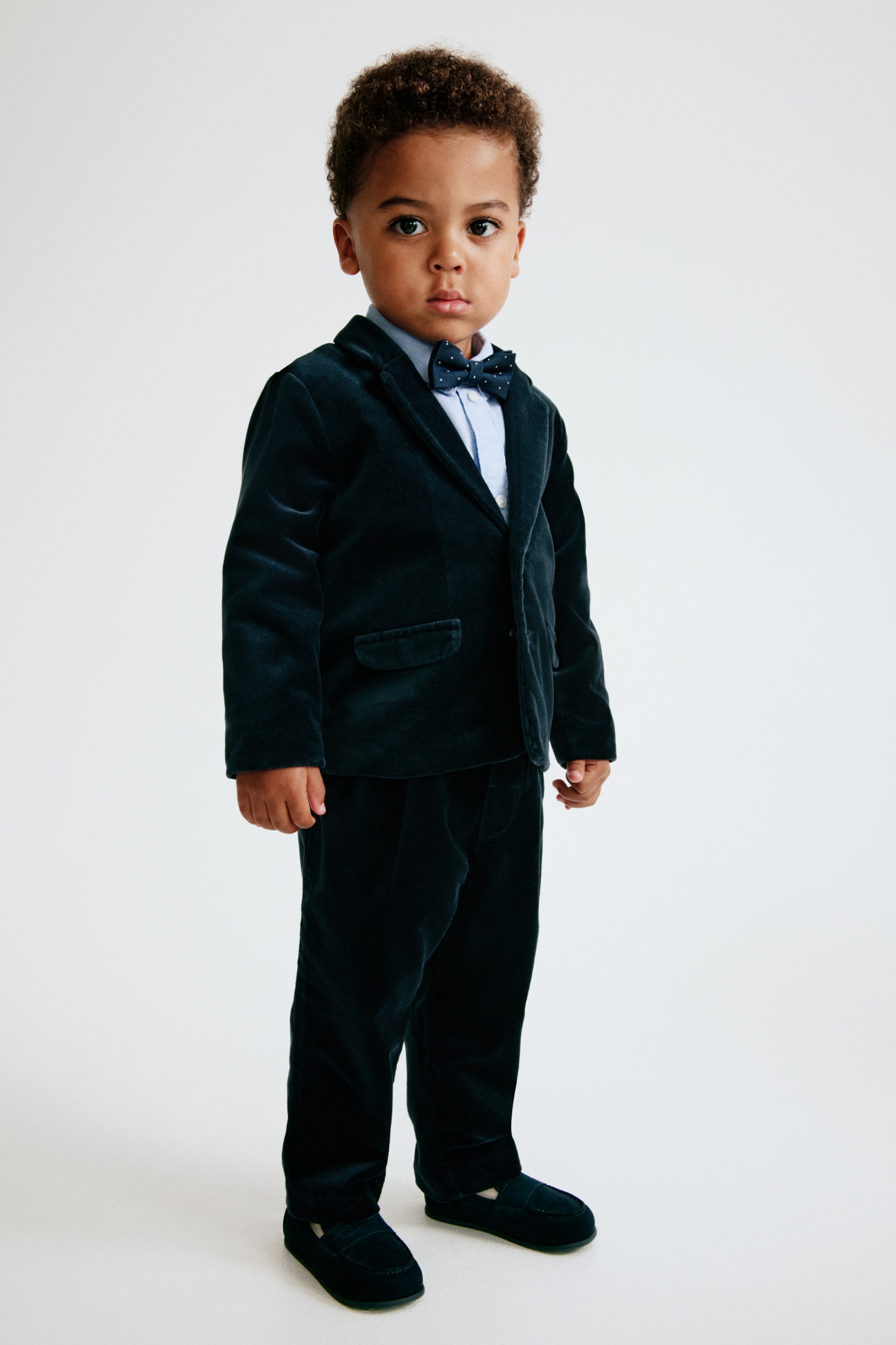 View larger image: This dressy set is shown from a three-quarter front angle, featuring a dark teal velvet jacket with notch lapels and matching velvet trousers. The jacket is layered over a light blue Oxford shirt and accessorized with a coordinating patterned bow tie.