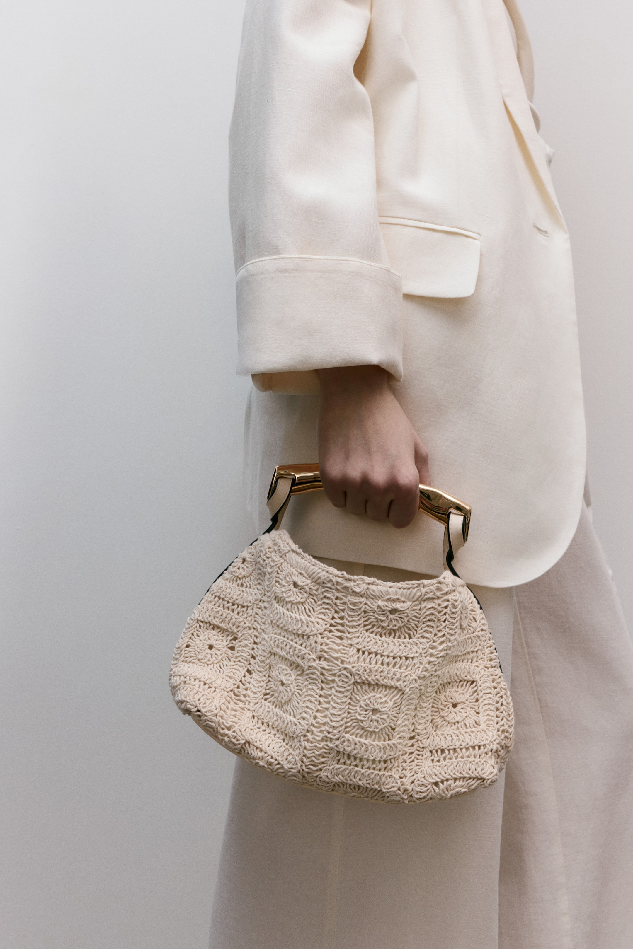 View larger image: The cream-coloured, crocheted handbag showcases square-like motifs with circular centres, and is held by a hand, highlighting its gold-toned, rigid top handles connected by thin dark straps.