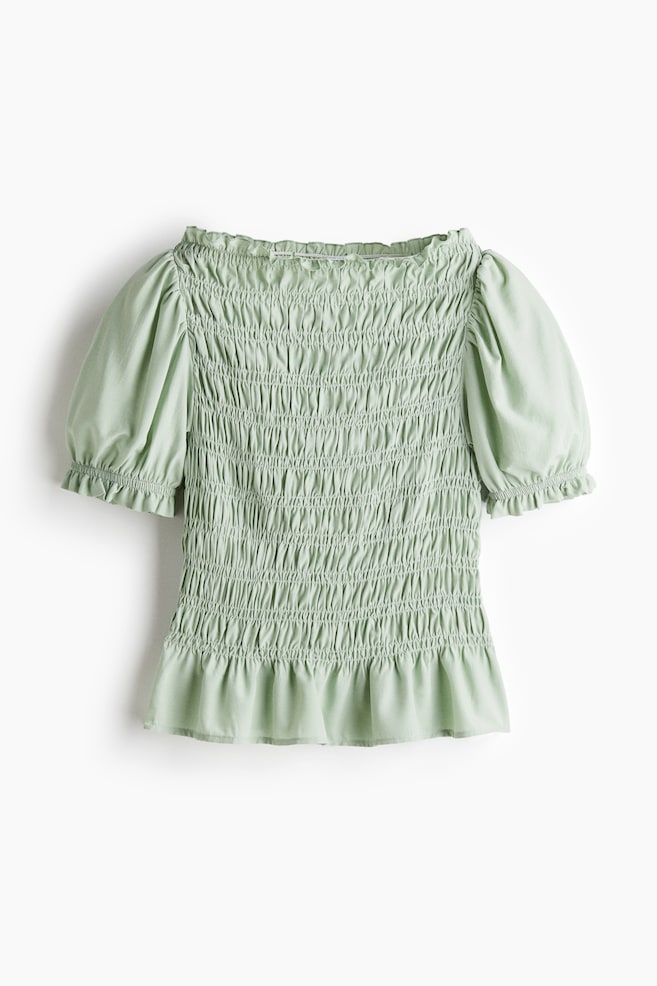 A light green blouse is displaying its FRONT, featuring a fitted body in a smocked weave. It is designed with a boat neckline, short puff sleeves with narrow elastication and a frill trim at the cuffs, and a flared hem. - 1