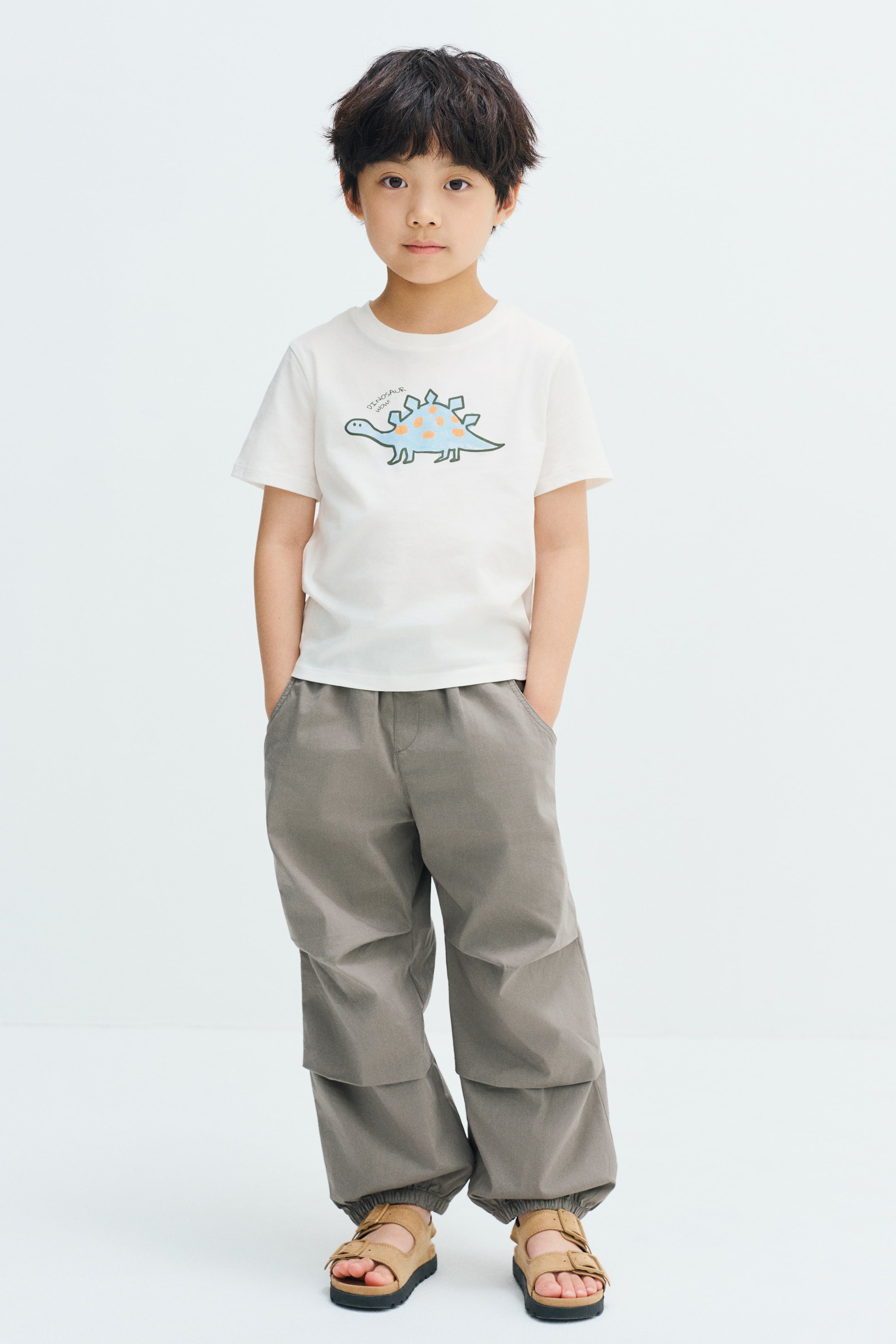 View larger image: A cream-coloured cotton jersey T-shirt featuring a printed dinosaur design on the chest and a round, rib-trimmed neckline is worn by the model. The T-shirt is styled with loose-fitting grey trousers and tan sandals.