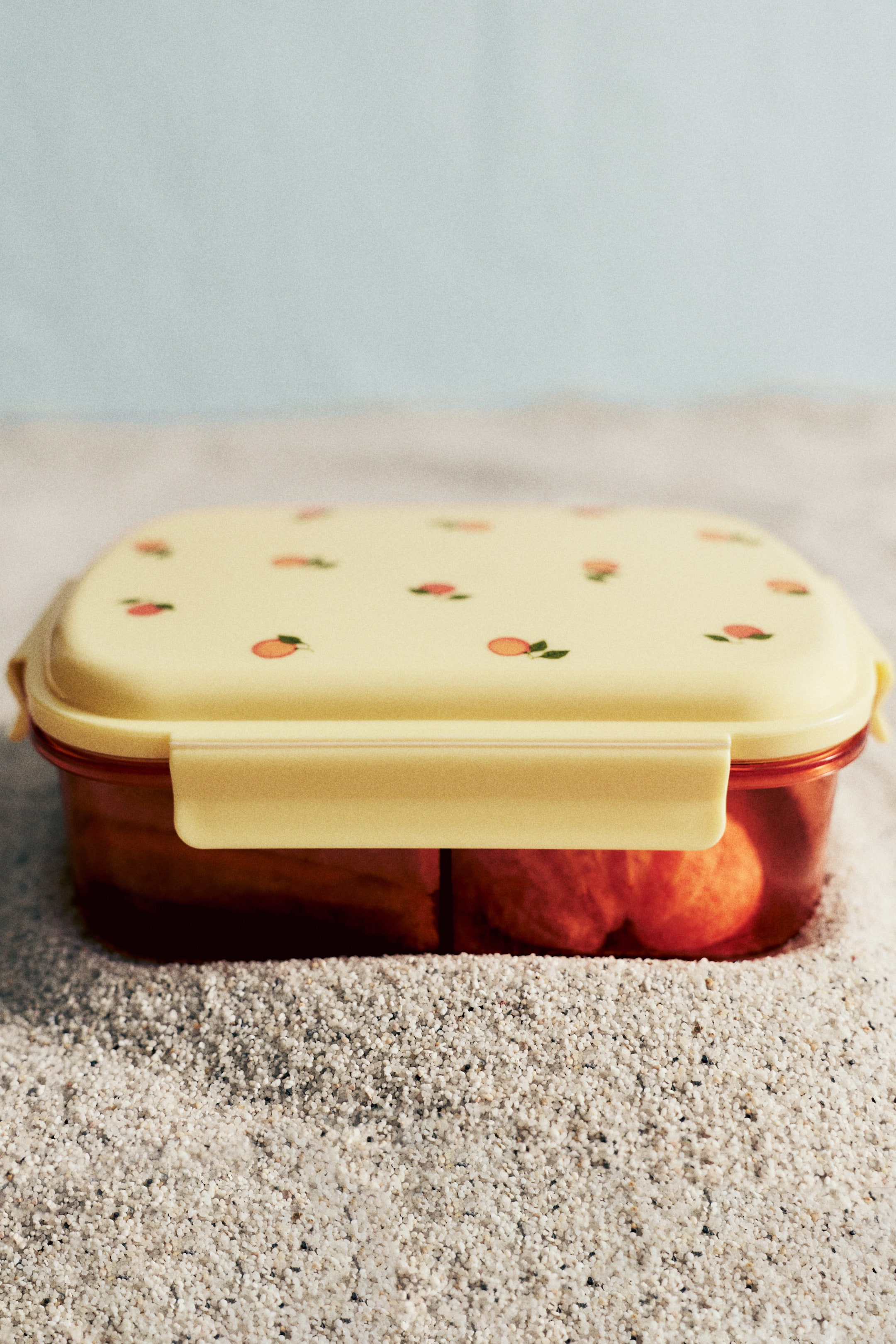 View larger image: A rectangular lunchbox is positioned facing forward, showcasing a pale yellow lid adorned with small peach illustrations and a translucent reddish-brown base. Pale yellow clips secure the lid, with reddish-orange food items visible inside the lunchbox.