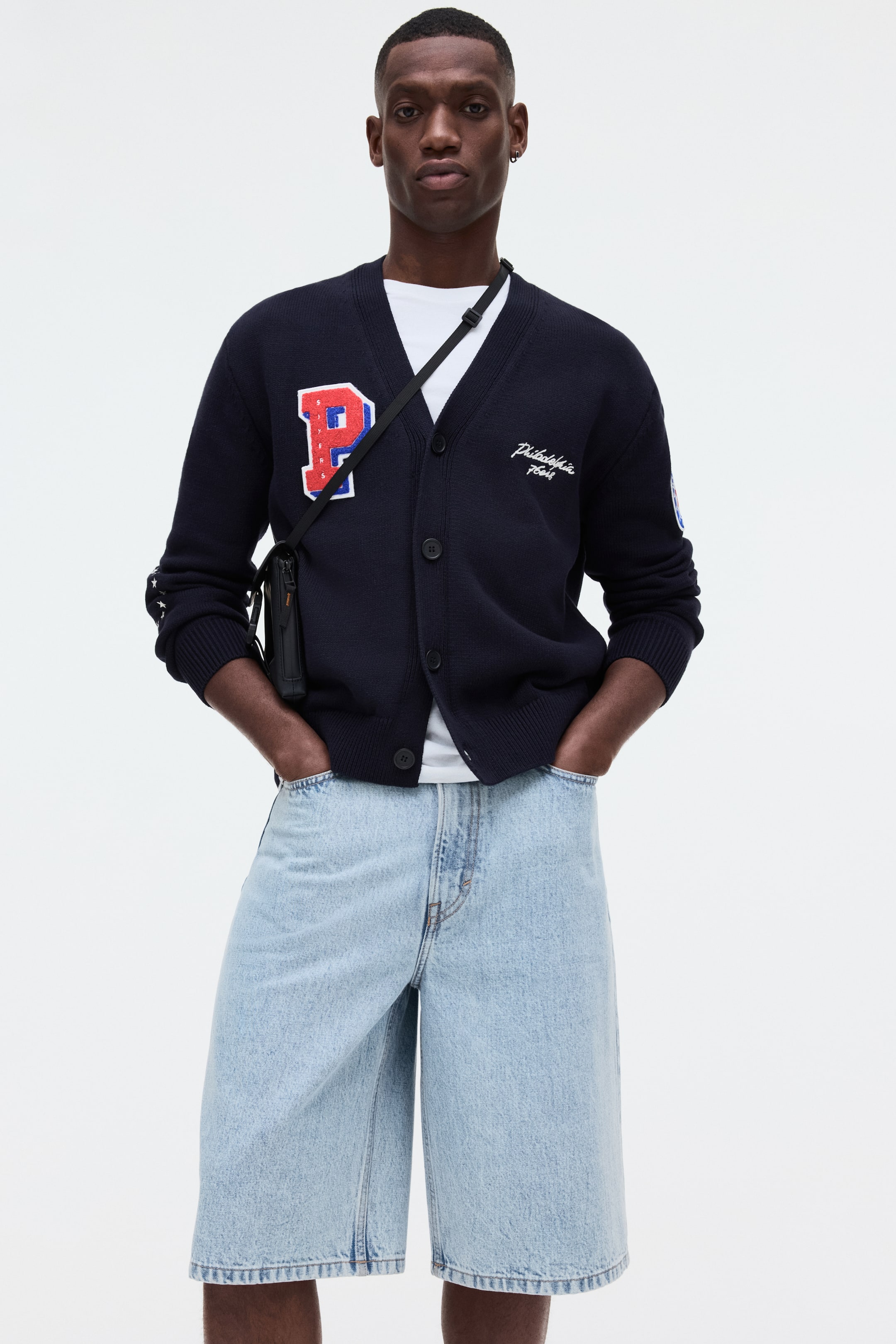 View larger image: A navy blue cardigan featuring a V-neckline, button-front closure with dark buttons, and ribbed cuffs and hem, is worn over a white t-shirt. A large red "P" patch with a white outline and blue stitching is visible on the left chest, while the word "Philadelphia" and a four-digit number are embroidered in white on the right chest.