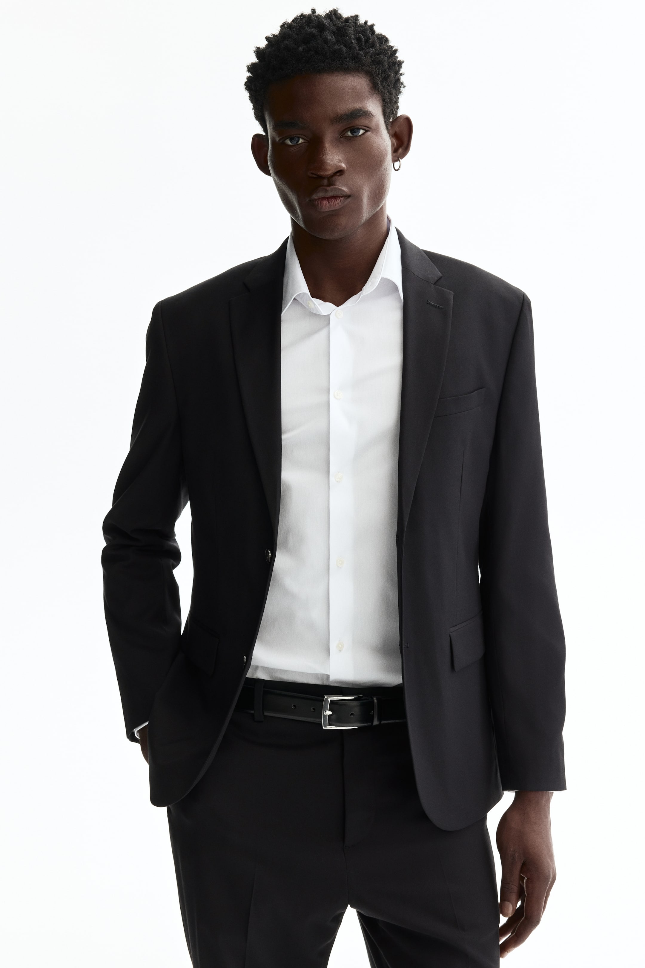 Agrandir l'image: Un modèle porte une veste noire à simple boutonnage, superposée à une chemise blanche à col. La veste présente des revers crantés étroits avec une boutonnière décorative, une poche poitrine passepoilée et des poches avant à rabat ; elle est boutonnée à l'avant et a une coupe ajustée, tandis que le modèle repose une main dans l'une de ses poches avant.