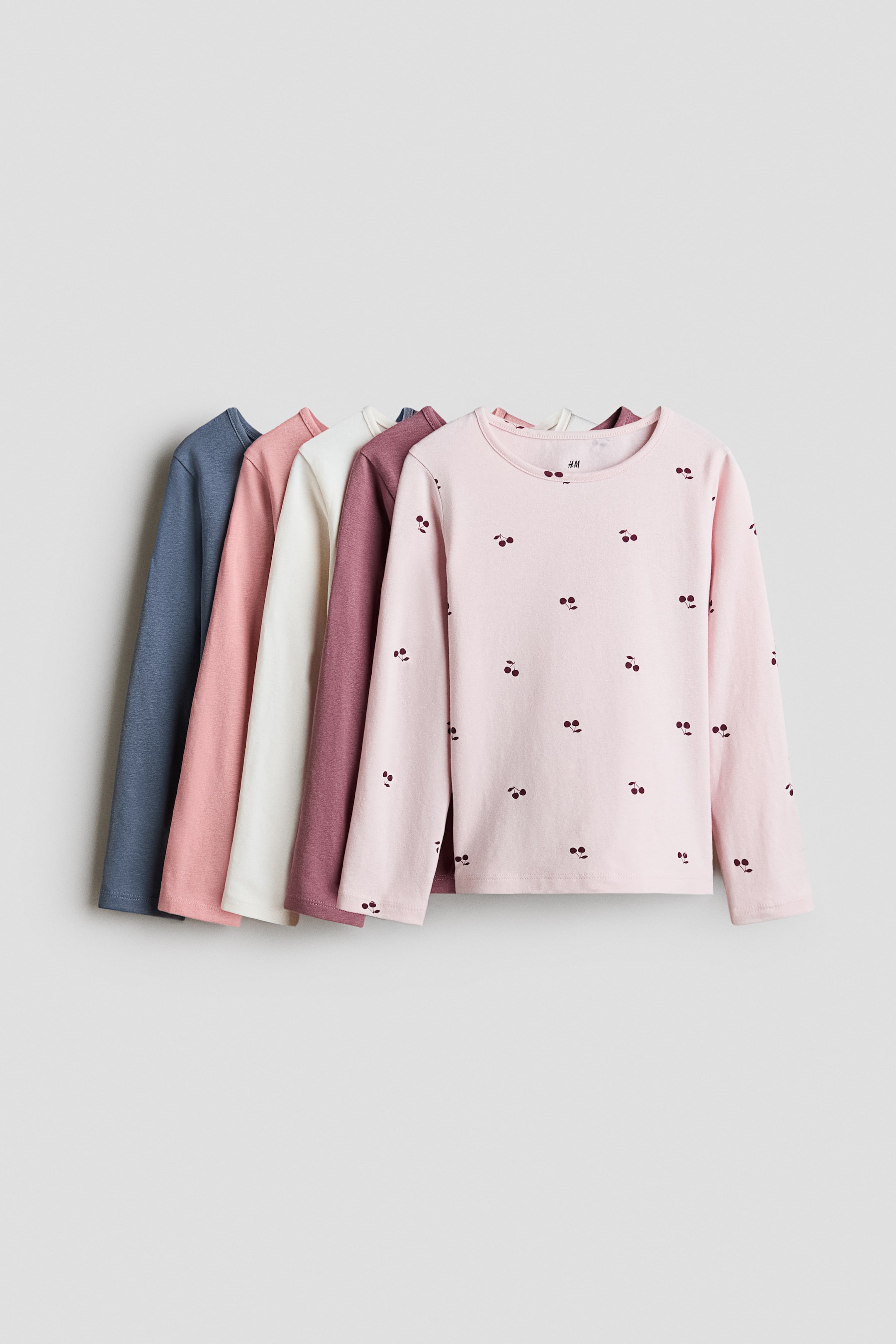 View larger image: Five long-sleeved tops are displayed in a layered arrangement, angled to the right. The front top features a light pink colour with a scattered dark cherry pattern, while the four tops behind it are in solid shades of mauve, cream, dusty pink, and dusty blue.