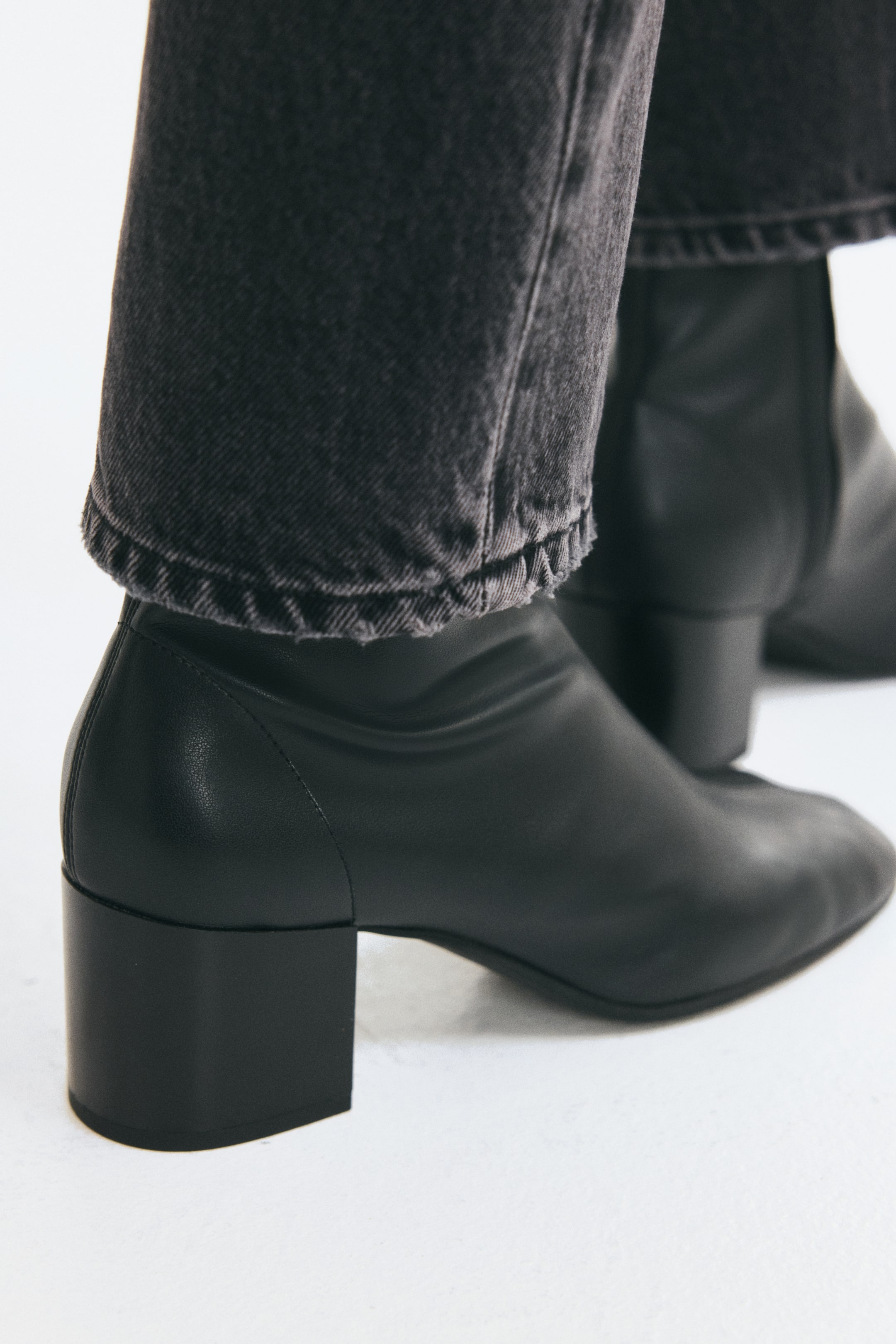 View larger image: Black block-heeled sock boots with rounded toes are being worn by the model. The boots feature a fitted shaft at the ankle, which is layered underneath the hem of dark grey denim jeans.