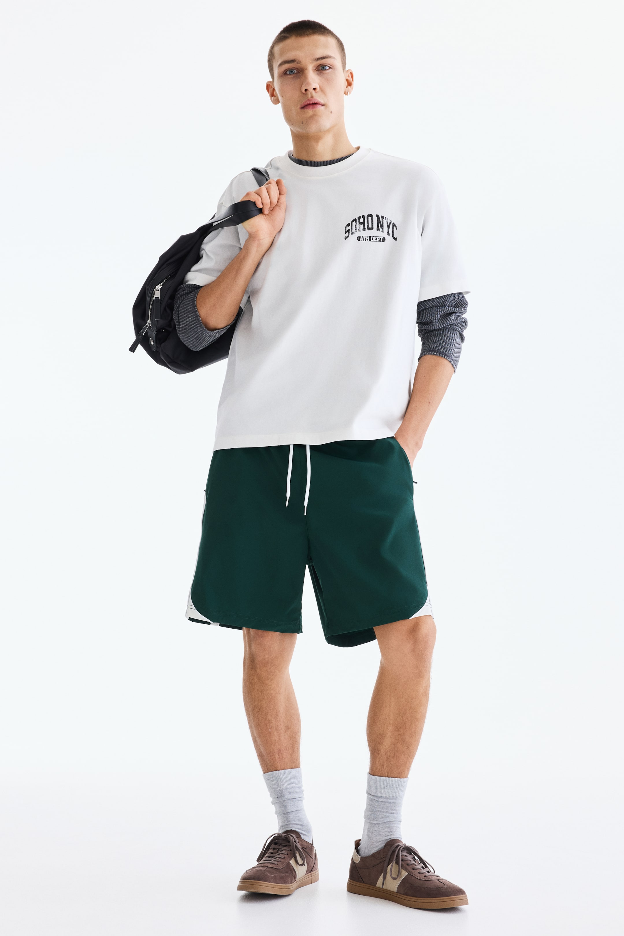 Ampliar la imagen: Una camiseta blanca de gran tamaño presenta un gráfico negro "SOHO NYC ATHL DEPT" en el pecho. Tiene cuello redondo y mangas cortas, con una camiseta de manga larga acanalada en gris visible debajo de sus mangas.