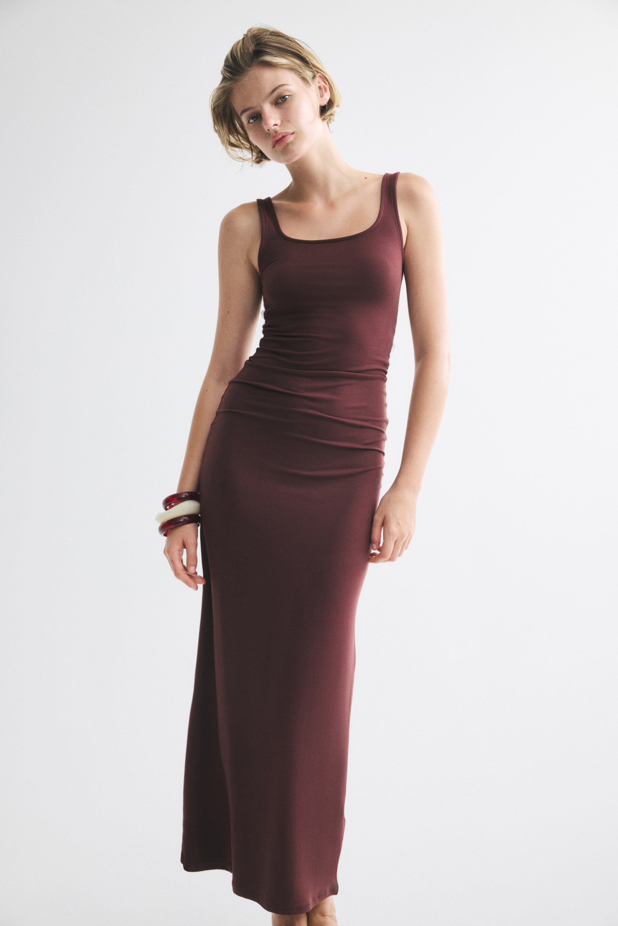View larger image: A long, fitted dress in dark burgundy stretch jersey is worn by the model, featuring a scoop neckline, wide shoulder straps, and a straight-cut hem. The dress is styled with thick, stacked bangles on the model’s wrist.