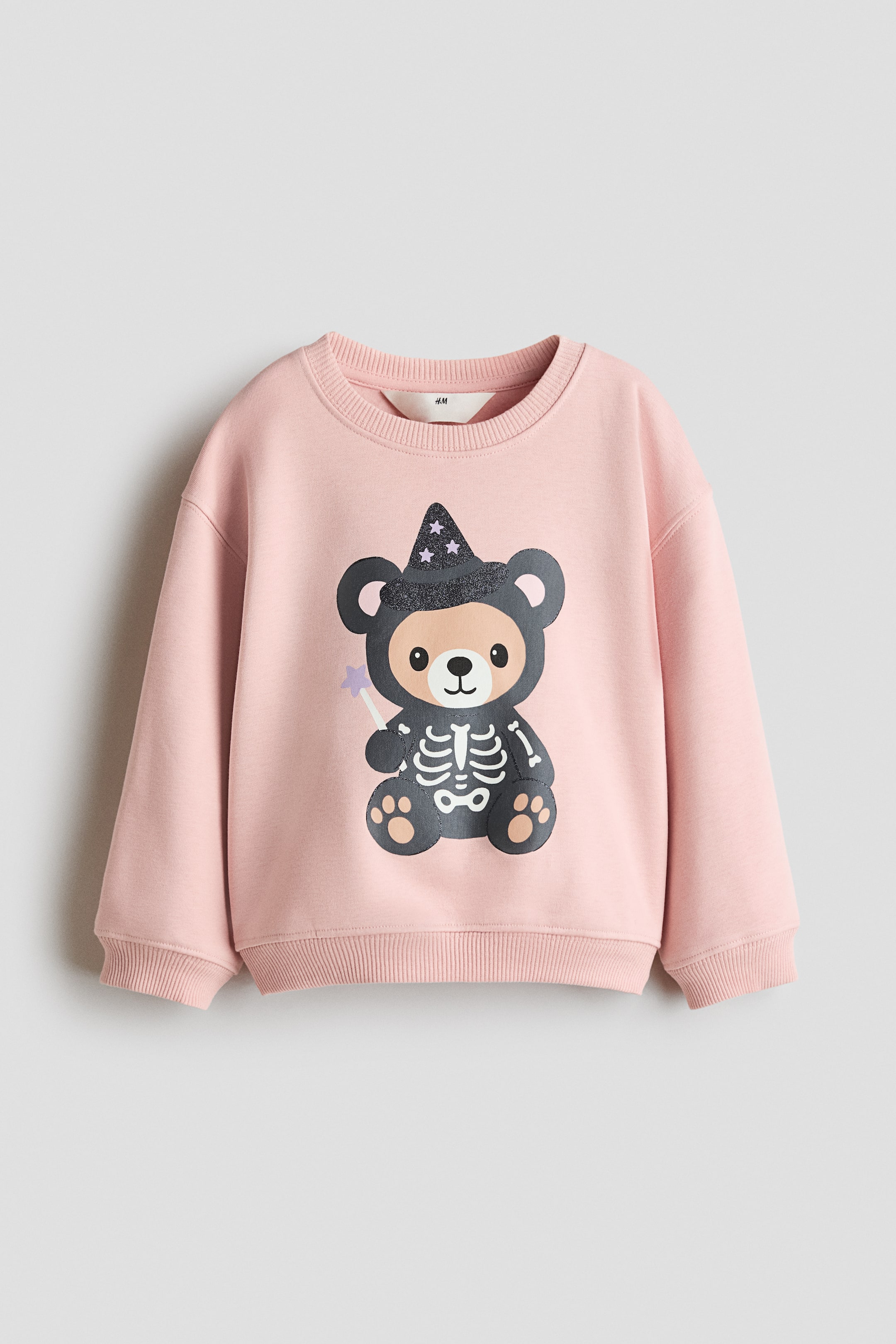 Größeres Bild ansehen: Ein hellrosa Sweatshirt ist von vorne zu sehen und verfügt über einen runden, mit Rippstrick eingefassten Halsausschnitt, überschnittene Schultern sowie Rippbündchen an den Ärmeln und am Saum. Auf der Vorderseite ist eine Grafik eines Cartoon-Bären zu sehen, der einen spitzen Hexenhut und ein Skelettkostüm trägt und einen Zauberstab mit Sternenspitze hält.