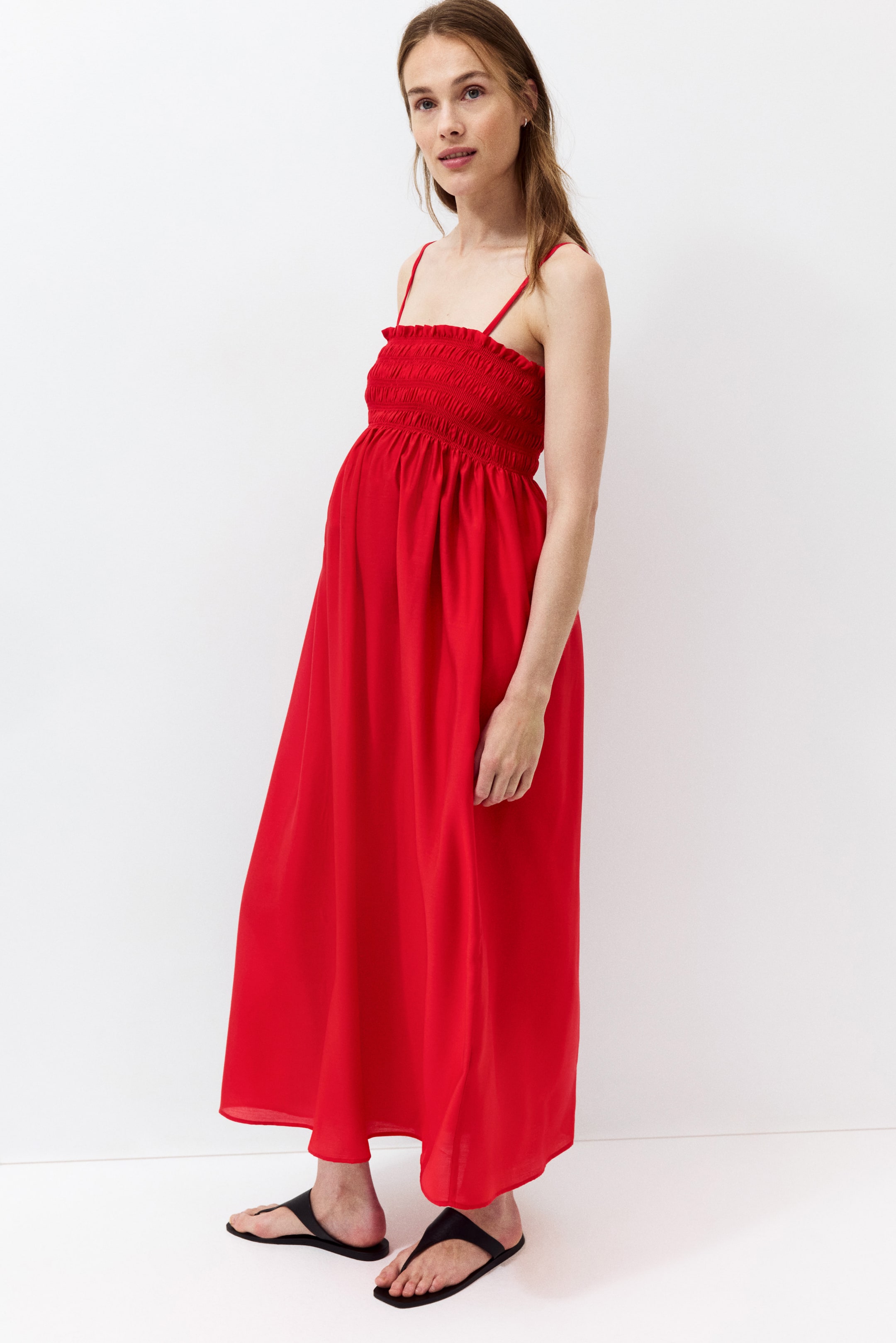 Größeres Bild ansehen: Rotes Maxikleid mit einem gesmokten Oberteil, das einen gerüschten oberen Rand und dünne Spaghettiträger aufweist. Das Kleid fällt fließend ab der gesmokten Taille, legt sich locker über den Bauch und reicht bis zu den Knöcheln. Es wird mit schwarzen flachen Sandalen gestylt.