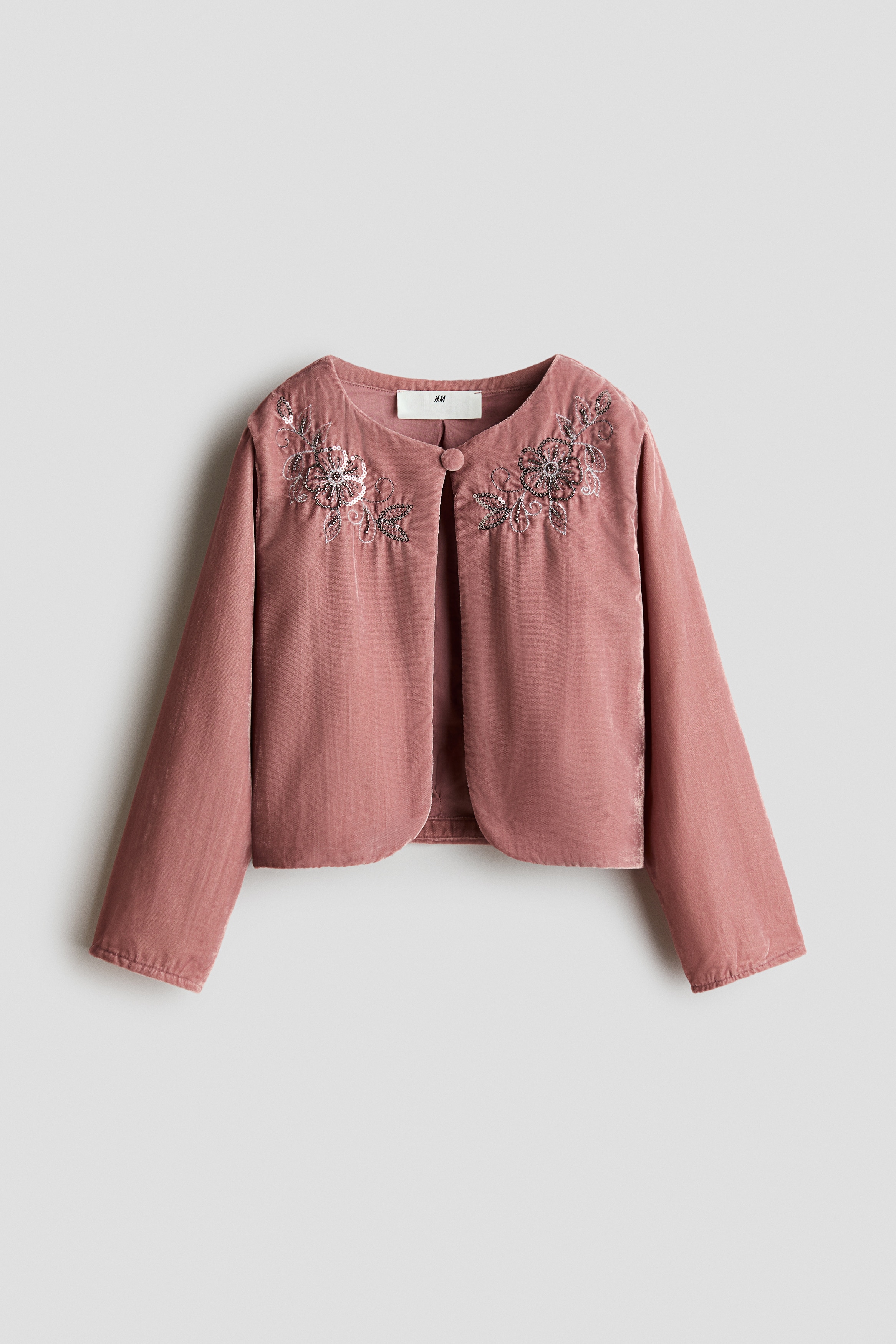 A dusty pink crinkled velvet jacket is shown from the front, featuring silver floral bead embroidery on the upper chest and a round neckline with a fastened covered button. The long-sleeved jacket falls open below the single-button fastening, worn over a white top and matching velvet trousers.