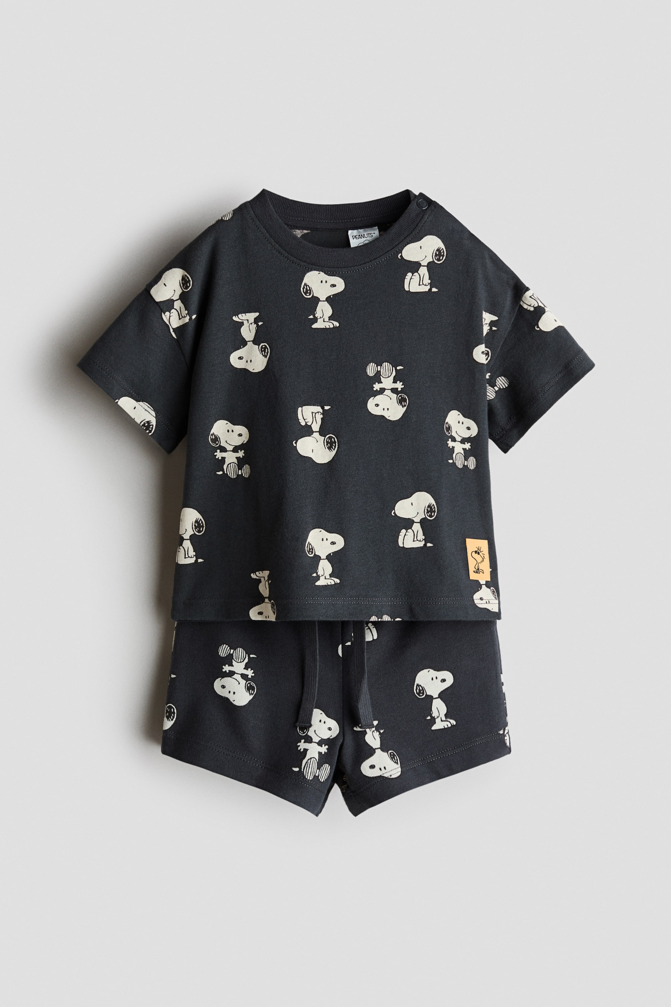 拡大画像を表示: 2点セットプリント。ダークグレーの半袖Tシャツは、クルーネックにスナップボタンが肩に付いており、全体にライトクリームのスヌーピープリントが施されています。コーディネートされたダークグレーのショーツは、同じスヌーピープリントとドローストリングウエストを特徴としており、どちらのアイテムも前面に提示されています。