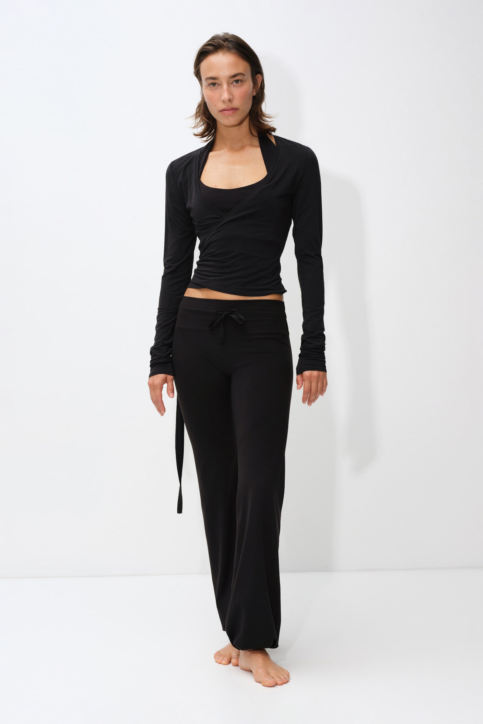 The model is wearing a black, slim-fit activewear top with long sleeves, a deep V-neckline, and a wrap front with ties hanging at the waist. The top is layered over a matching black tank top and styled with black wide-leg trousers. - 1