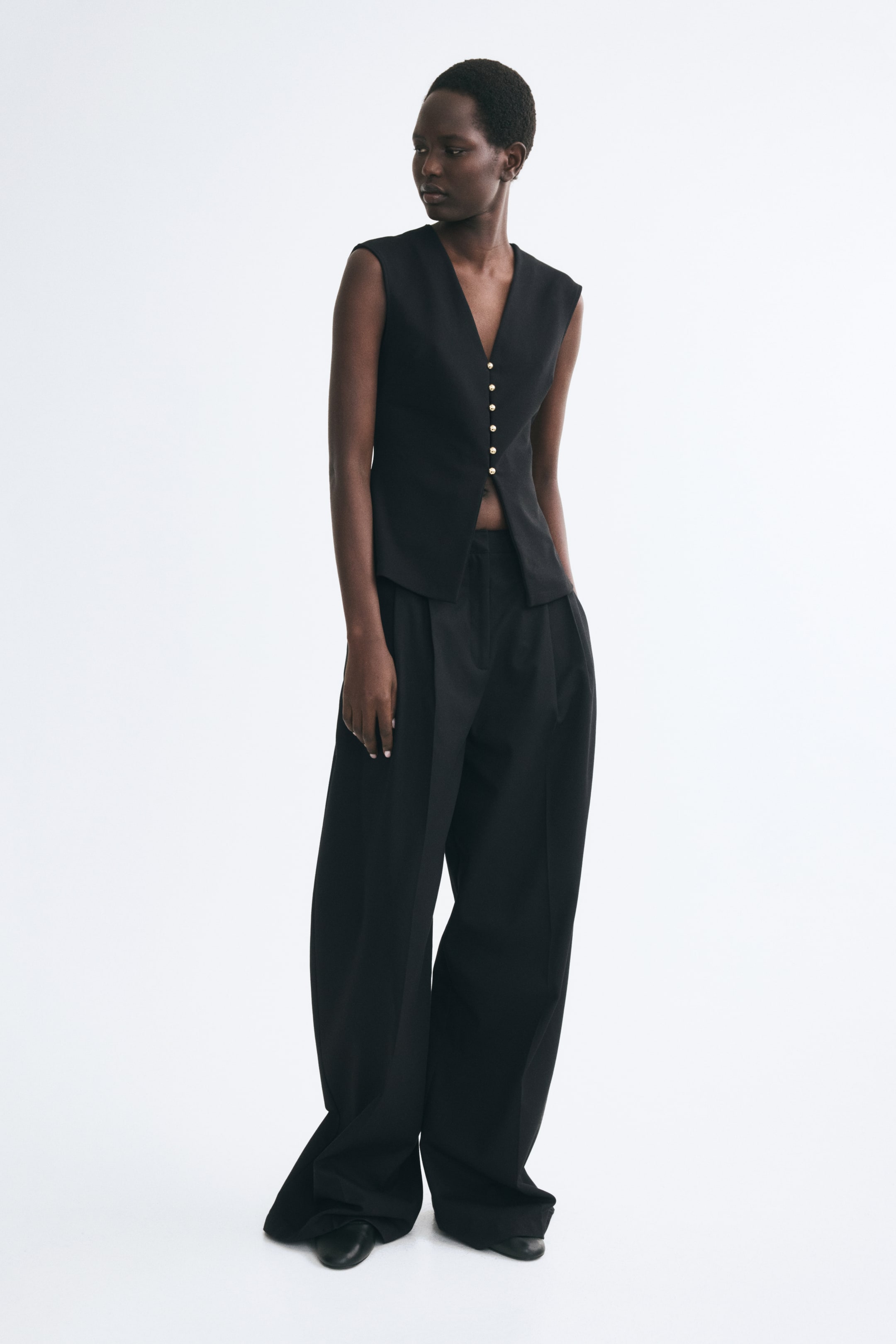 Ampliar la imagen: La modelo lleva un chaleco negro de tejido ponte con un escote pronunciado en forma de V y botones metálicos abombados al frente. El chaleco sin mangas se combina con pantalones negros de pierna ancha a juego.