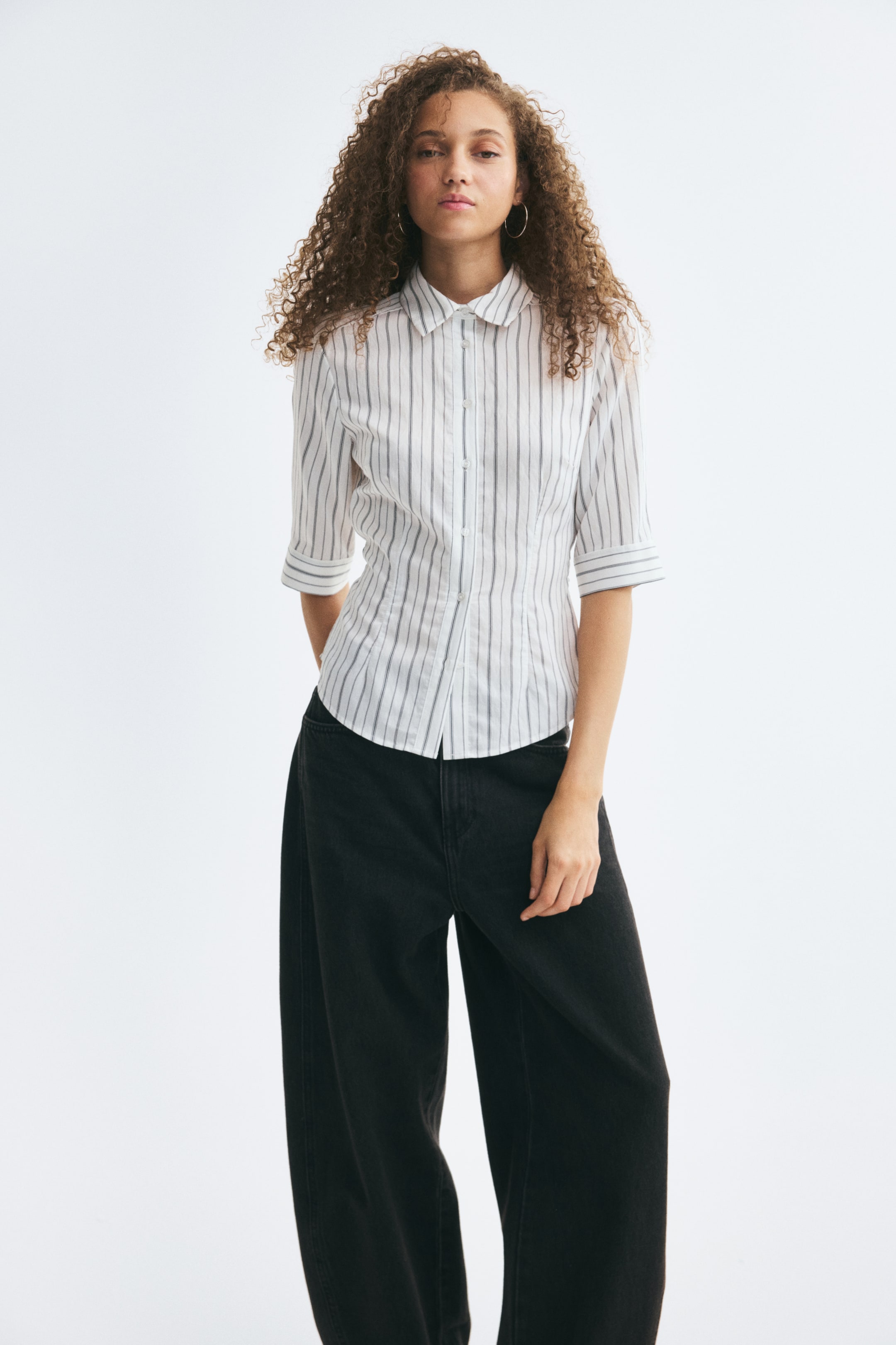 View larger image: The model is wearing a fitted white shirt with vertical black stripes, featuring a collar, button-down front, 3/4-length sleeves, and waist darts for a tailored silhouette. The shirt has a rounded hem and is styled with black wide-leg trousers.
