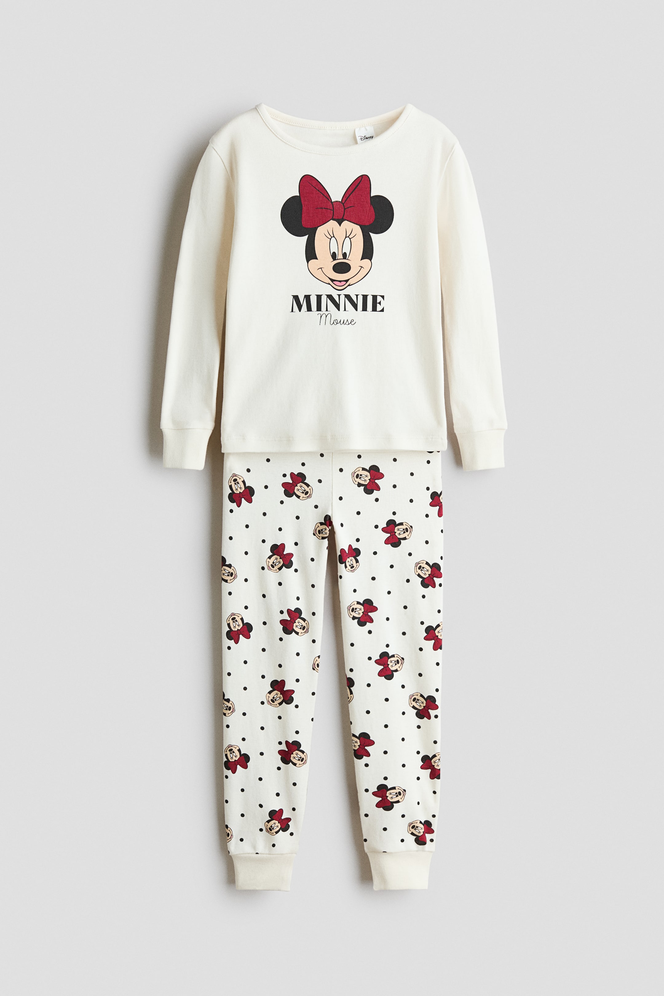 View larger image: A cream-coloured pyjama set is shown in a front view, featuring a long-sleeve top and matching pants. The top has a round neckline with narrow trim, ribbed cuffs, and a large Minnie Mouse graphic printed on the chest, while the pants feature an all-over print of Minnie Mouse heads and black polka dots with ribbed hems.