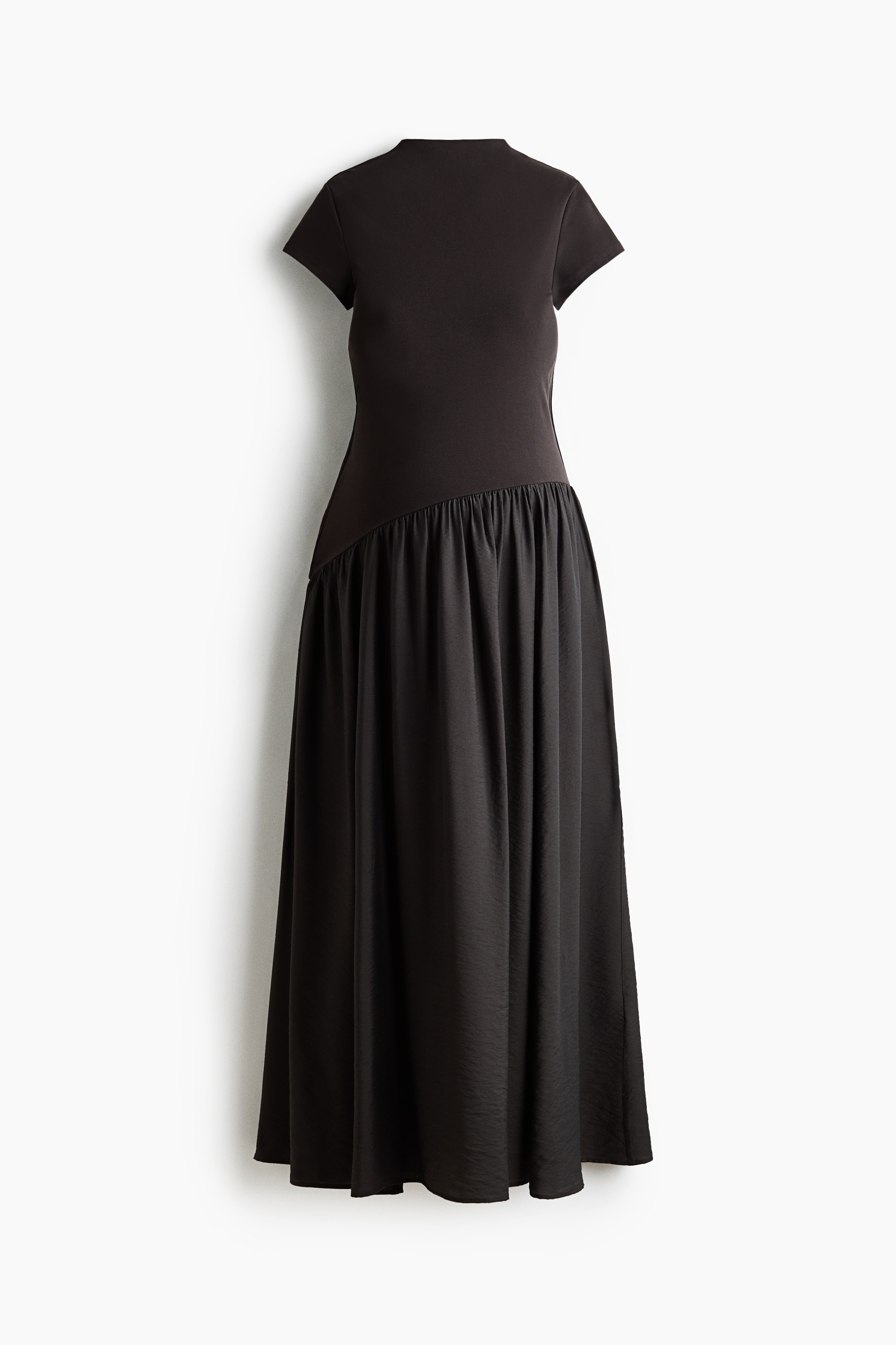 A dark brown maxi dress is worn by the model, featuring a stand collar, cap sleeves, a fitted bodice, and an asymmetric gathered seam at the hips that flows into a flared skirt. The dress is styled with light-coloured, pointed-toe heeled shoes as the model stands facing forward.