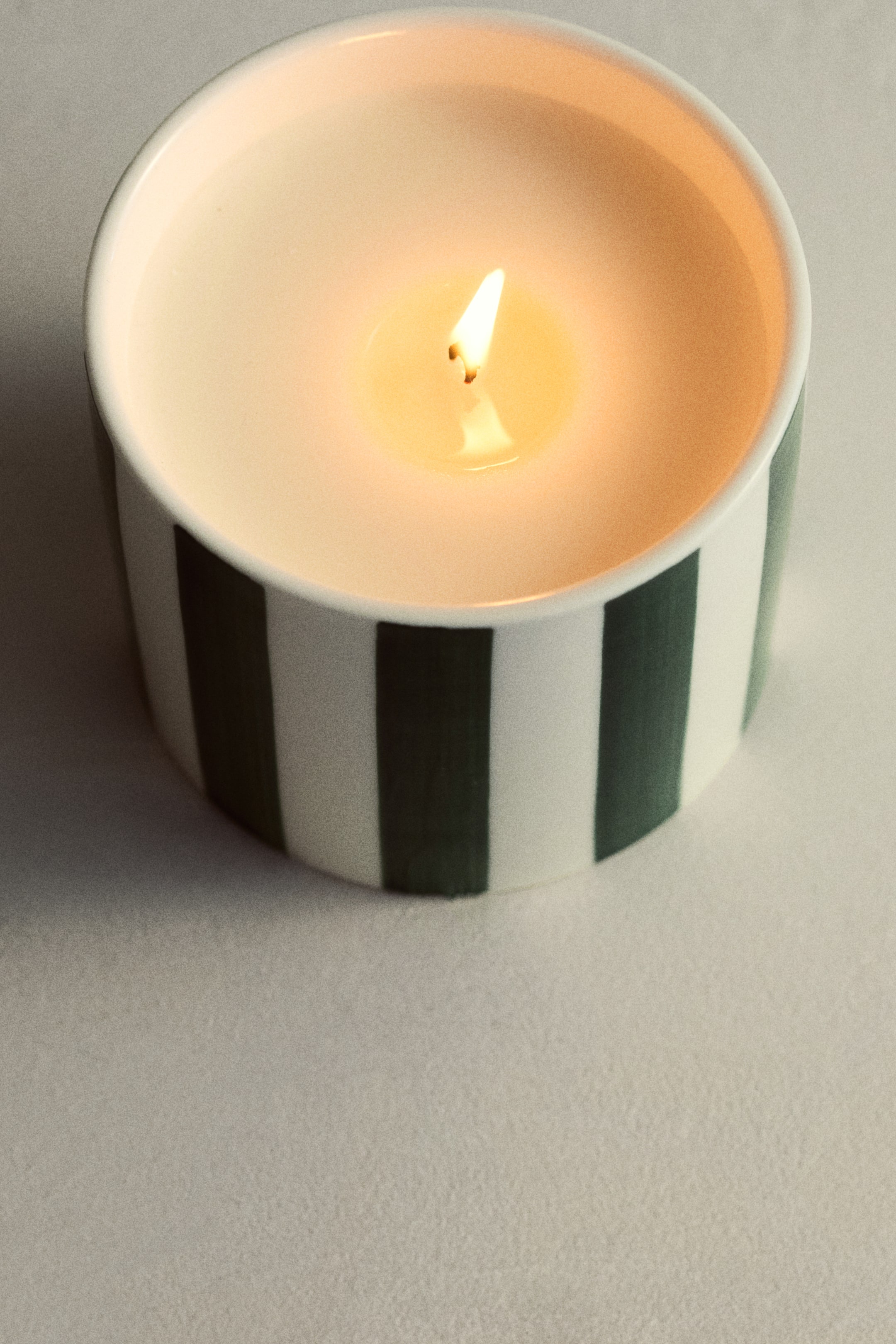 Grotere afbeelding bekijken: Een lichtgekleurde kaars brandt in een ronde keramische houder, met verticale donker groene en witte strepen.
