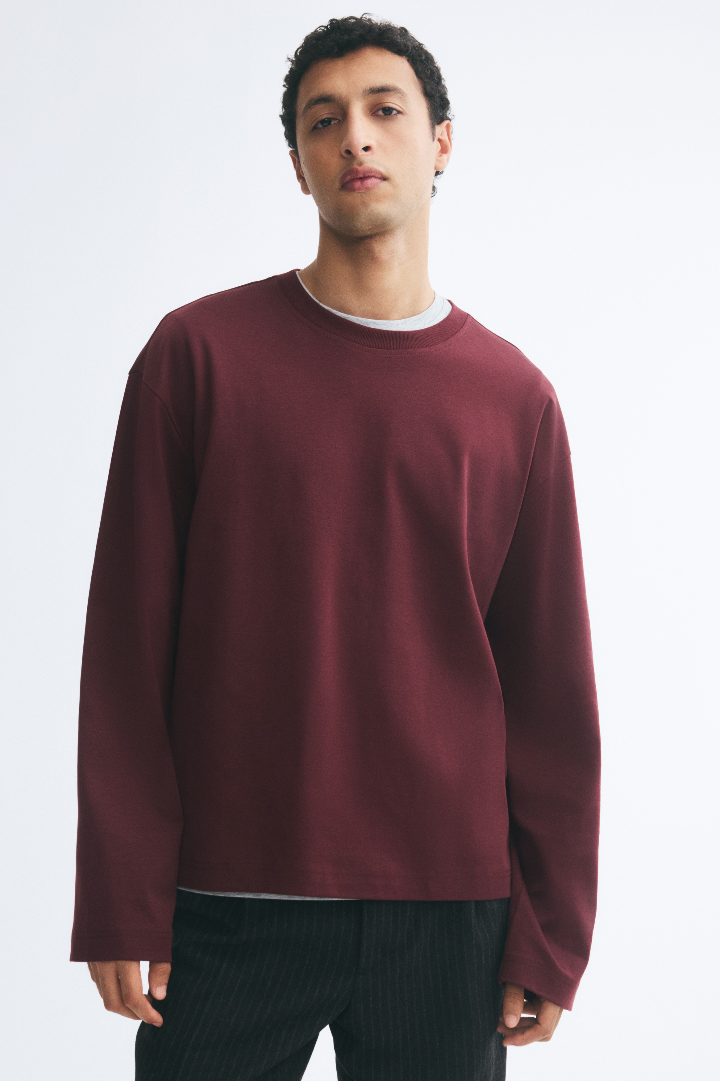 A burgundy long-sleeve T-shirt is hanging front-facing, showcasing a relaxed-fit silhouette with dropped shoulders and wide sleeves. The T-shirt features a ribbed crew neckline and a straight-cut hem.