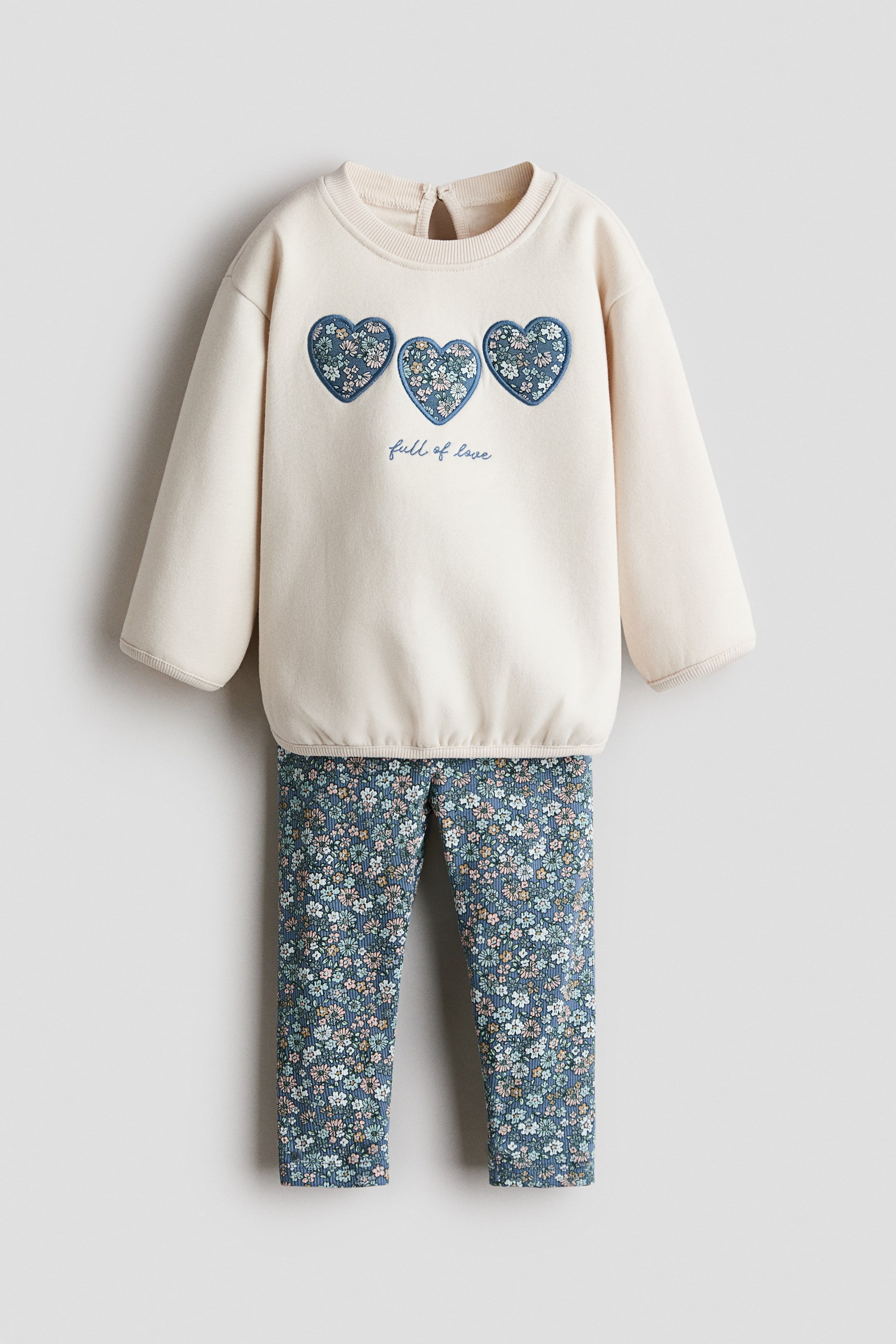 View larger image: A two-piece children's clothing set featuring an off-white sweatshirt with three heart-shaped blue floral patches and "full of love" embroidered on the front, paired with matching blue floral print leggings. The sweatshirt is designed with a ribbed crew neck, ribbed cuffs, and a ribbed hem, while the leggings feature a subtle ribbed texture.