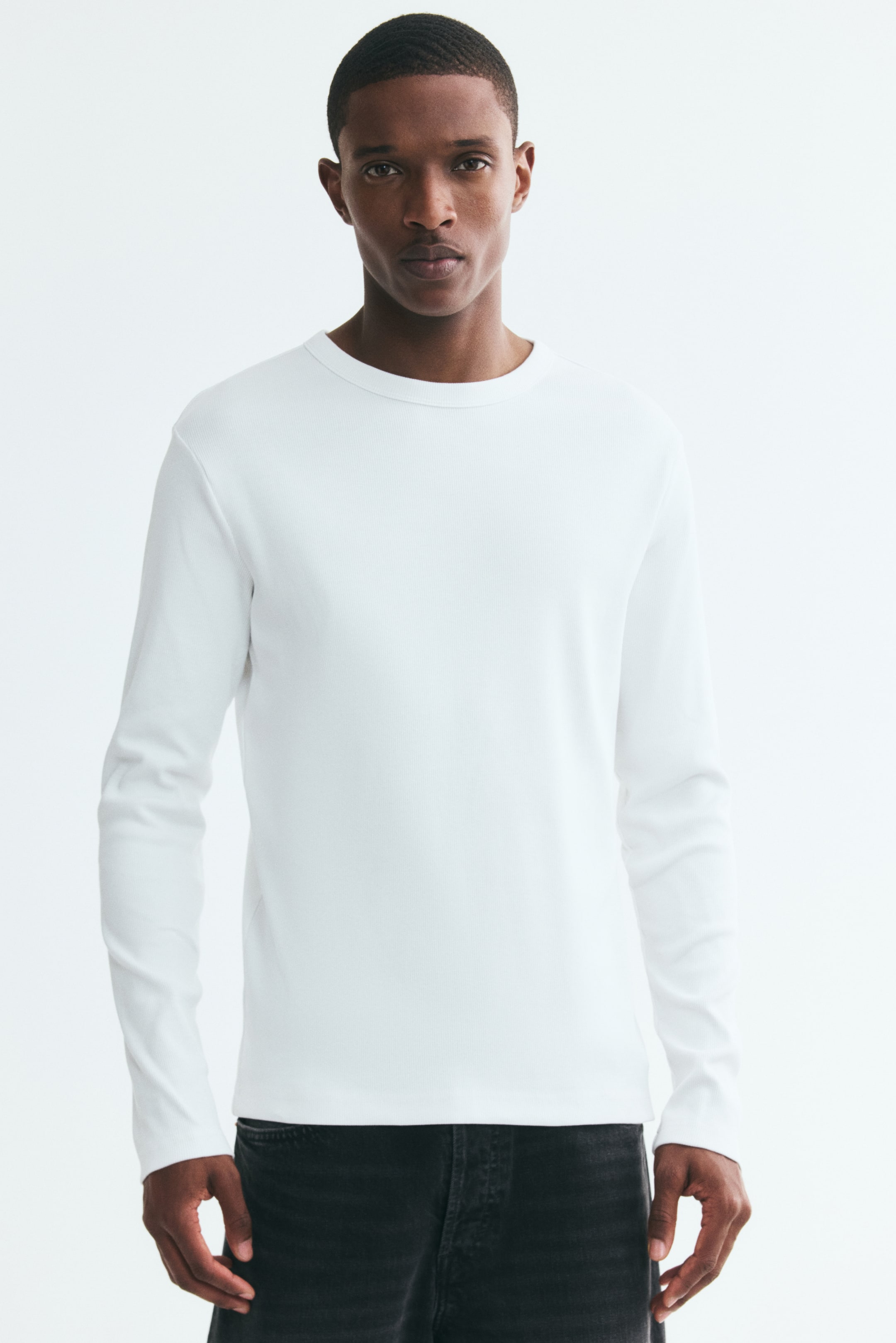 Büyük resmi görüntüle: Modelin üzerinde, yuvarlak yakasını, dar kalıbını ve düz kesimli etek ucunu öne çıkaran, fitilli pamuk jarse kumaştan beyaz, uzun kollu bir T-shirt giyilmektedir. T-shirt, koyu renk kot pantolon ile kombinlenmiştir.