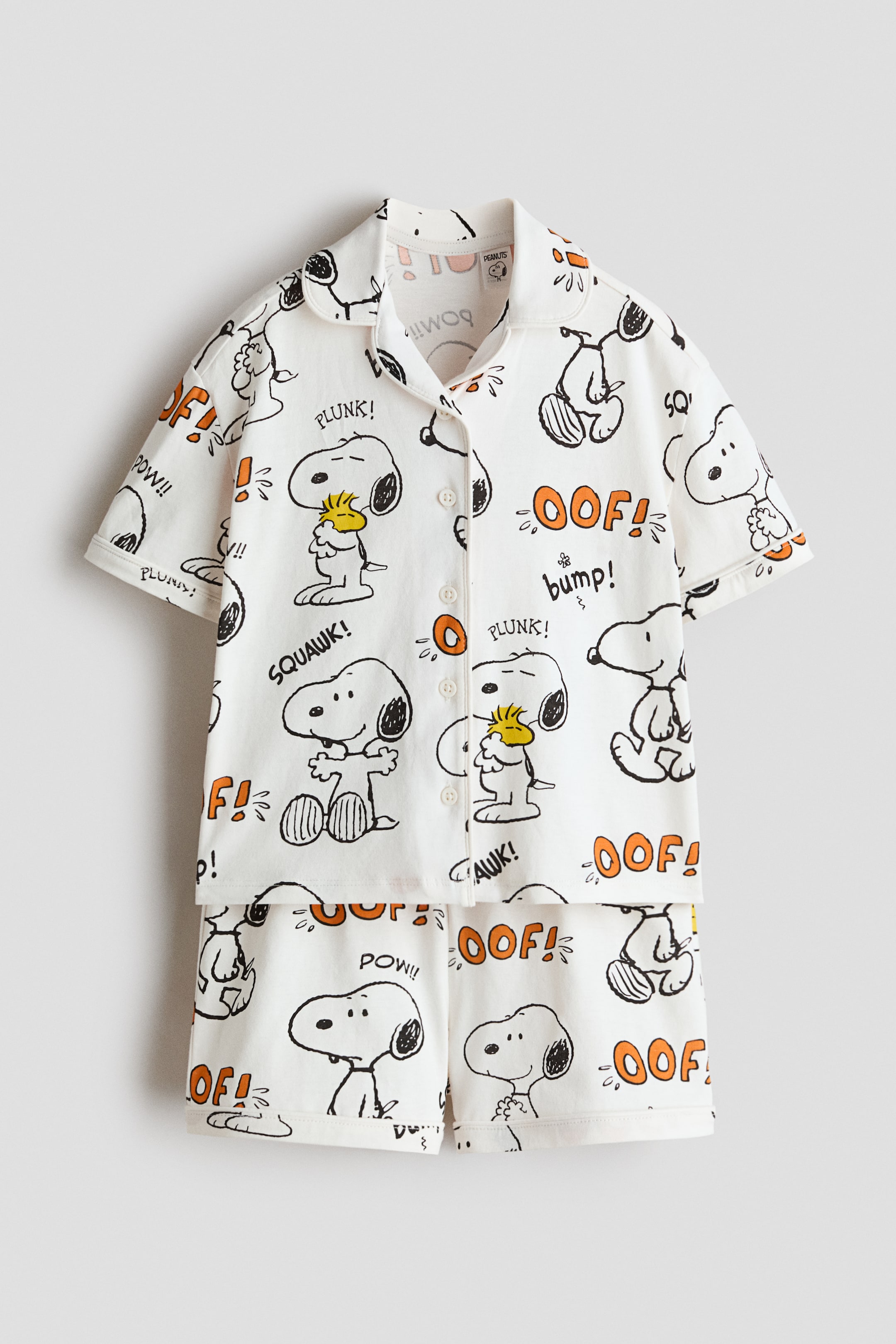 View larger image: A white two-piece pajama set is showcased front-facing, featuring a short-sleeved top with a revere collar and a button-up front, paired with matching shorts. Both pieces are adorned with a repeating pattern of Snoopy and Woodstock characters along with orange comic sound effect words like "OOF!" and "PLUNK!".