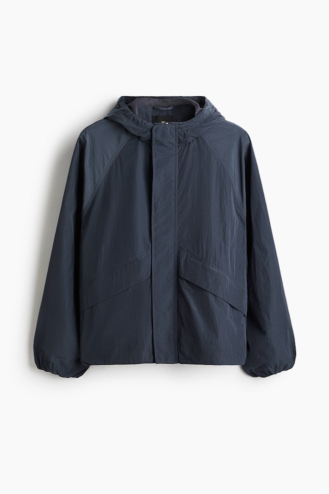 Um casaco leve de cor azul escuro com uma textura enrugada é mostrado de frente, apresentando um capuz com um forro de malha visível no interior. Também é desenhado com um fecho oculto, dois bolsos com abas inclinadas, mangas raglan e punhos elásticos. - 1