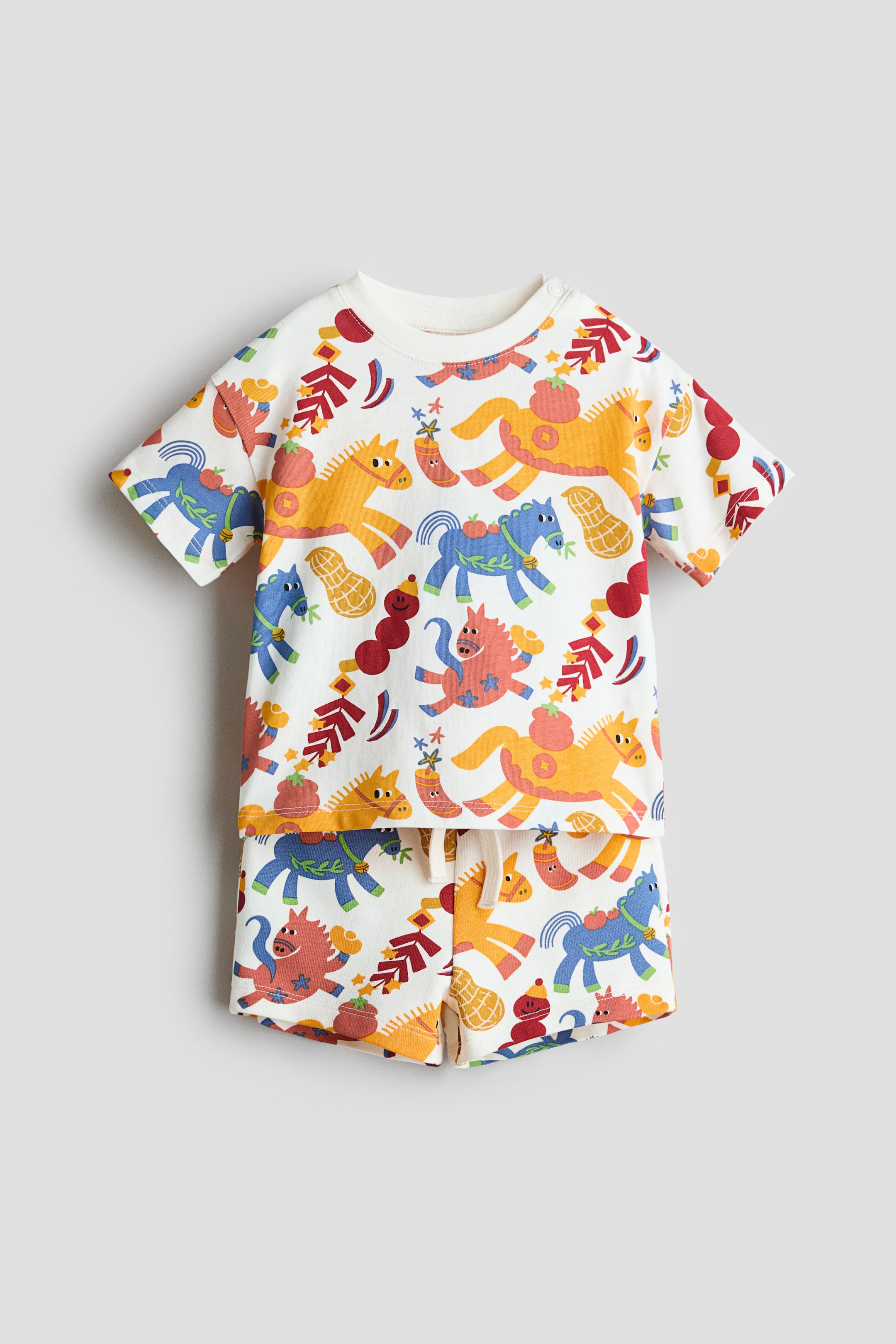 拡大画像を表示: 白い乳児用Tシャツとショーツのセットが平らに置かれ、カラフルなカートゥーンの馬、ピーナッツ、祝祭のモチーフが描かれています。Tシャツはショートスリーブとリブのクルーネックが特徴で、左肩に2つのスナップが付いており、マッチするショーツにはホワイトのドローストリング付きの伸縮性ウエストバンドが見られます。