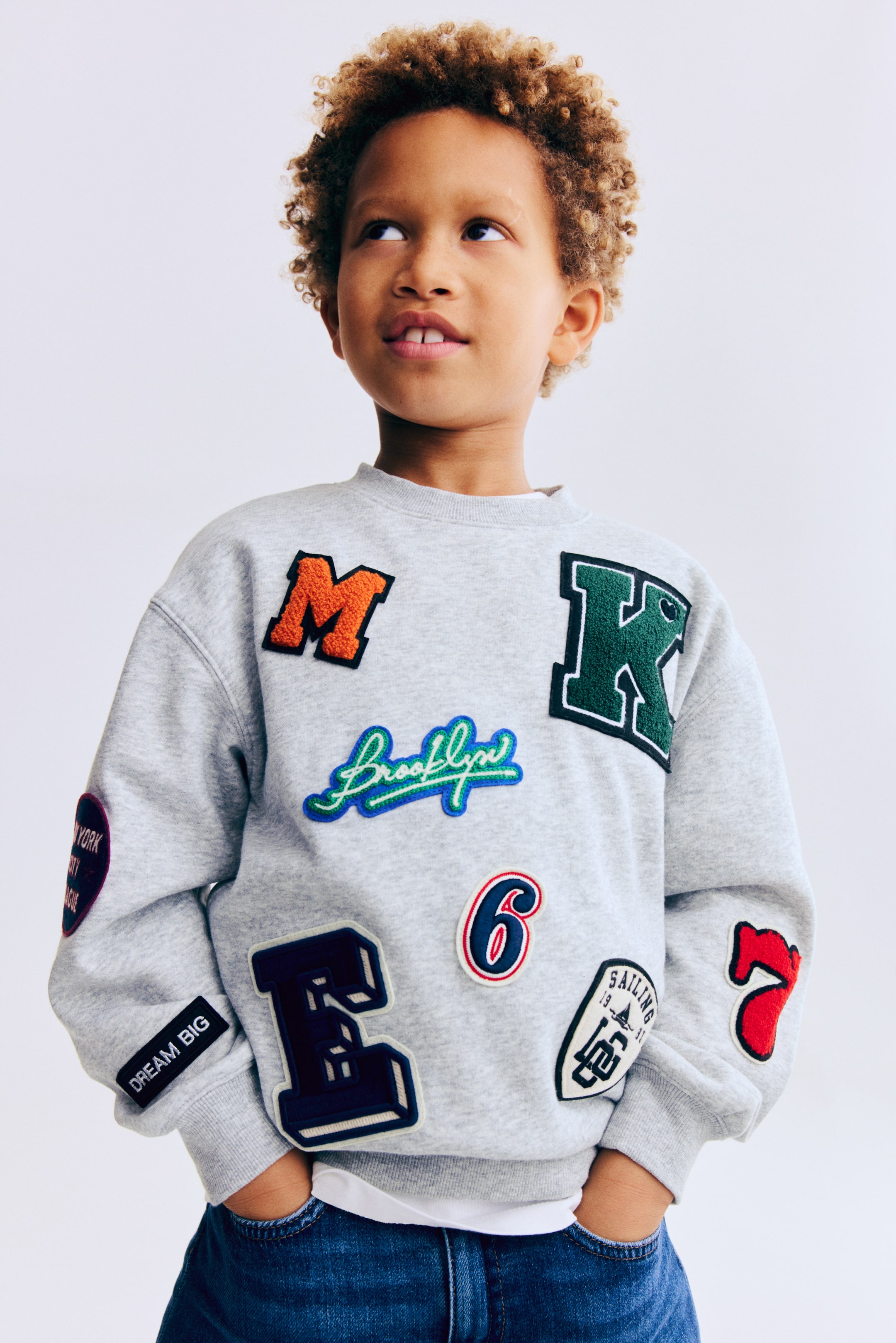 Agrandir l'image: Un sweat-shirt à col ras-du-cou gris clair présente de nombreux patchs brodés colorés, dont un grand 'M' orange, un 'K' vert foncé et un lettrage 'Brooklyn' bleu et vert, répartis sur le devant et les manches. Il est doté de bords-côtes aux poignets et à l'ourlet, et est porté par-dessus un t-shirt blanc et un jean bleu, présenté en vue avant légèrement oblique.