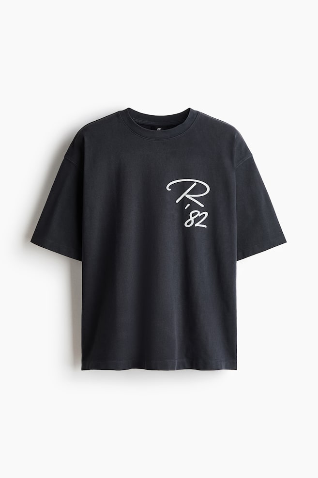 The front view of a washed black oversized t-shirt is displayed, highlighting its crew neck and short sleeves. The chest showcases a white stylized 'R.' with '82' below it. - 1