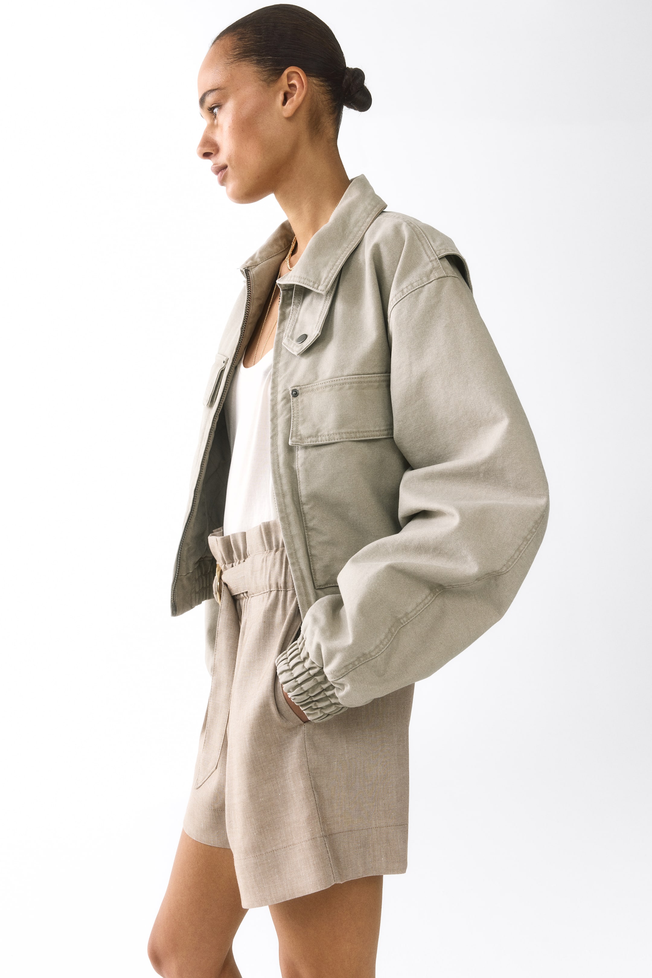 Größeres Bild ansehen: Leicht taupegrüne, gekürzte Utility-Jacke mit spitzem Kragen, einem teilweise geöffneten Frontreißverschluss und zwei Brusttaschen mit Klappe. Sie präsentiert lange Ärmel mit elastischen Bündchen und einen elastischen Saum, von der Seite betrachtet, offen über einem weißen Tanktop getragen.