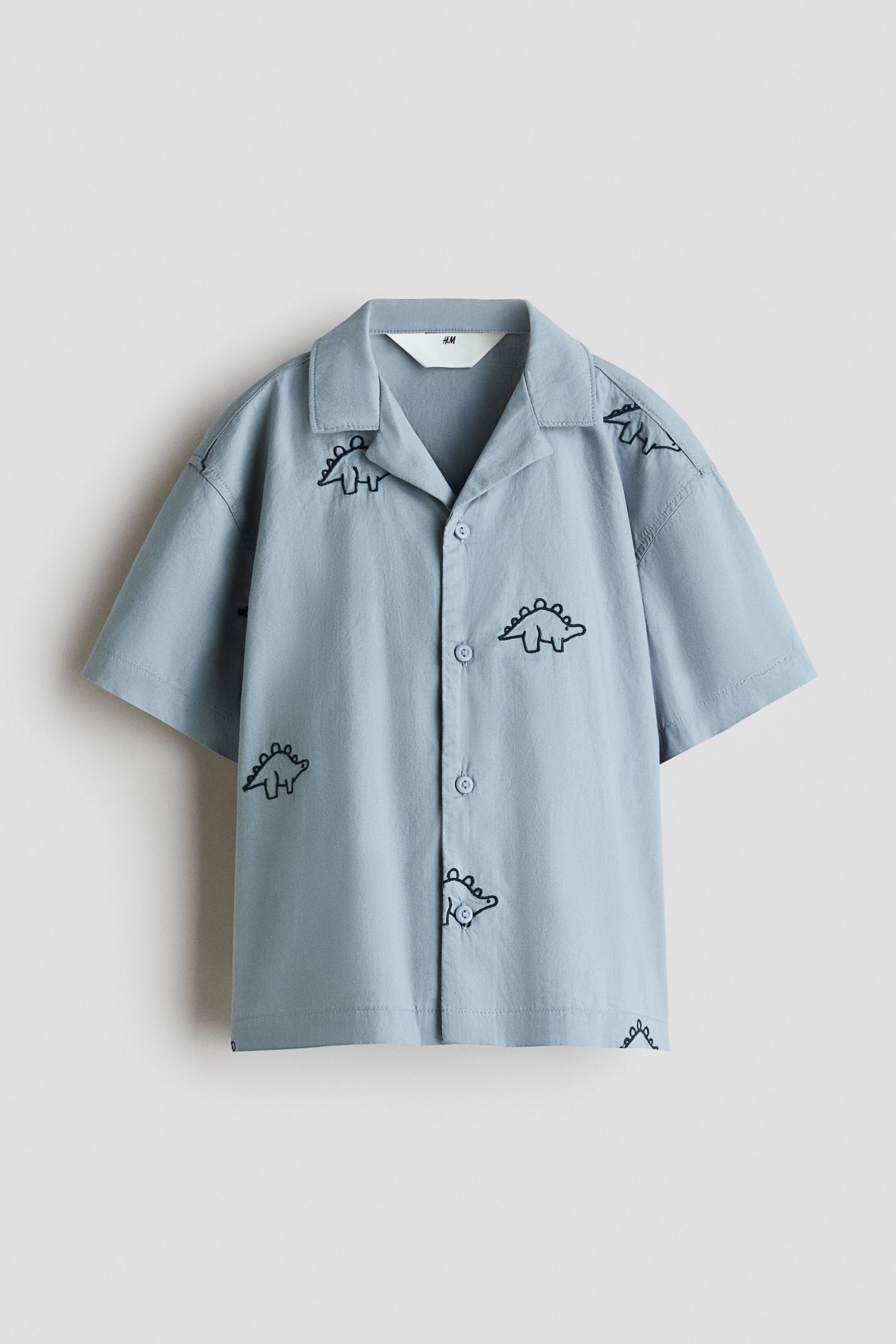 View larger image: A light blue short-sleeve shirt is shown from the front, featuring a Cuban collar and a button-up placket. Black outline dinosaur illustrations are patterned across the fabric.