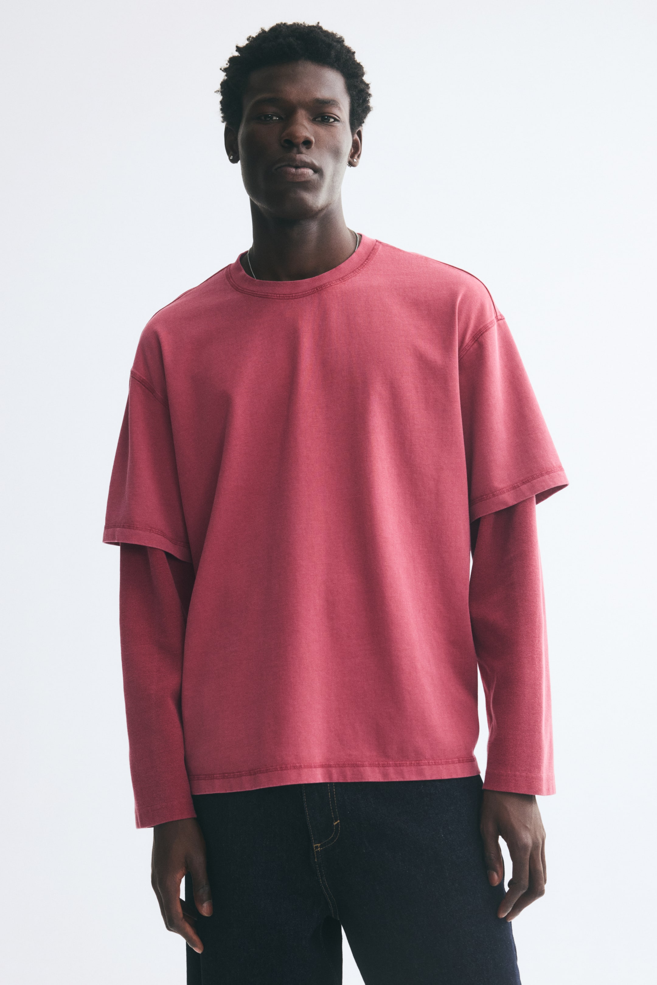 View larger image: The model is wearing a pink, relaxed-fit T-shirt featuring a layered design with long sleeves, dropped shoulders, and a round neckline. The heavyweight cotton jersey tee is styled with dark denim jeans.