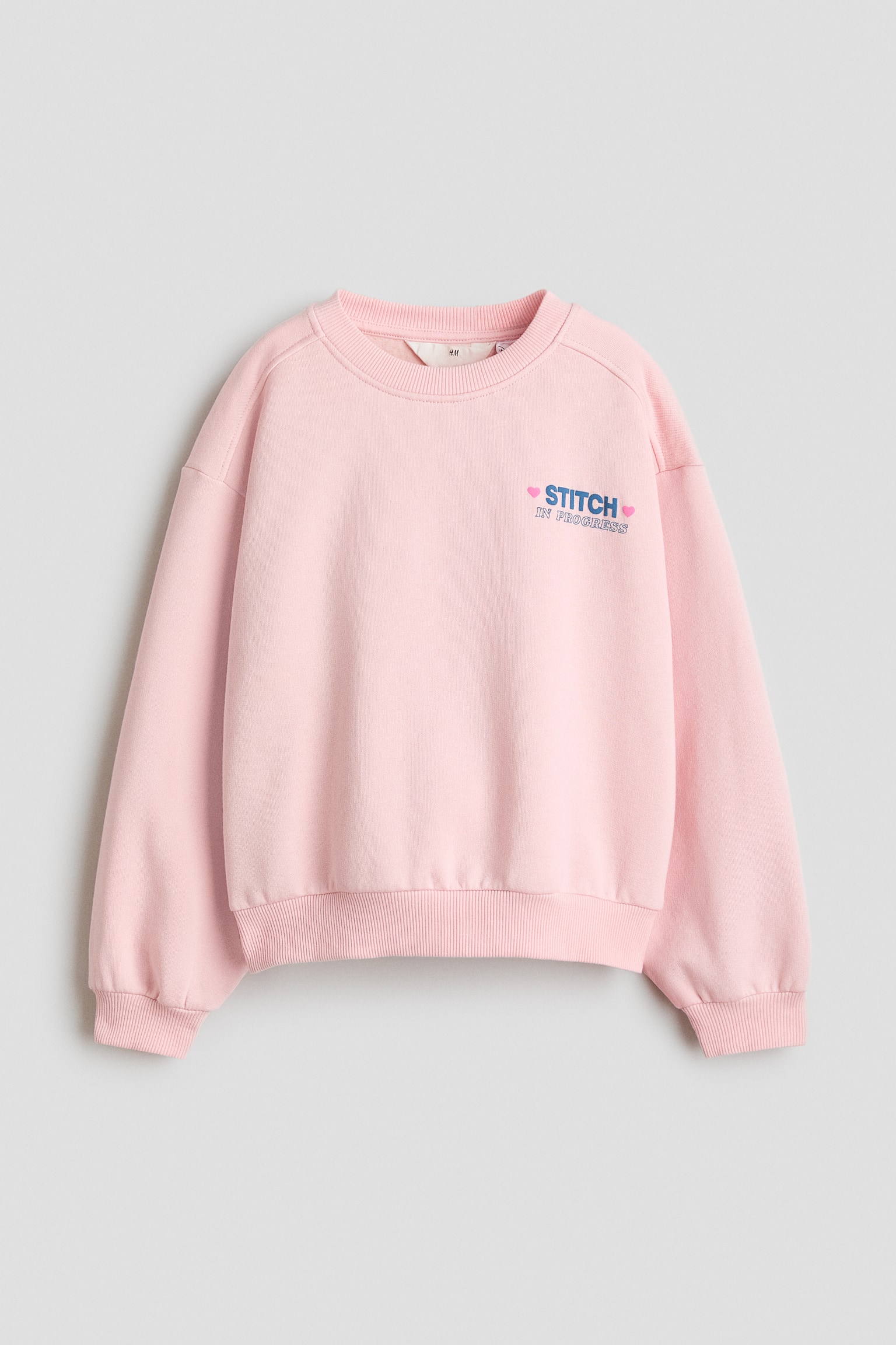 Uma camisola de fleece rosa-claro estÃ¡ virada para a frente, destacando um decote redondo com acabamento canelado, ombros descaÃ­dos e punhos e bainha canelados. A camisola apresenta um pequeno grÃ¡fico no peito do lado esquerdo com a inscriÃ§Ã£o "STITCH" em azul e "IN PROGRESS" em rosa, ladeado por dois pequenos coraÃ§Ãµes.