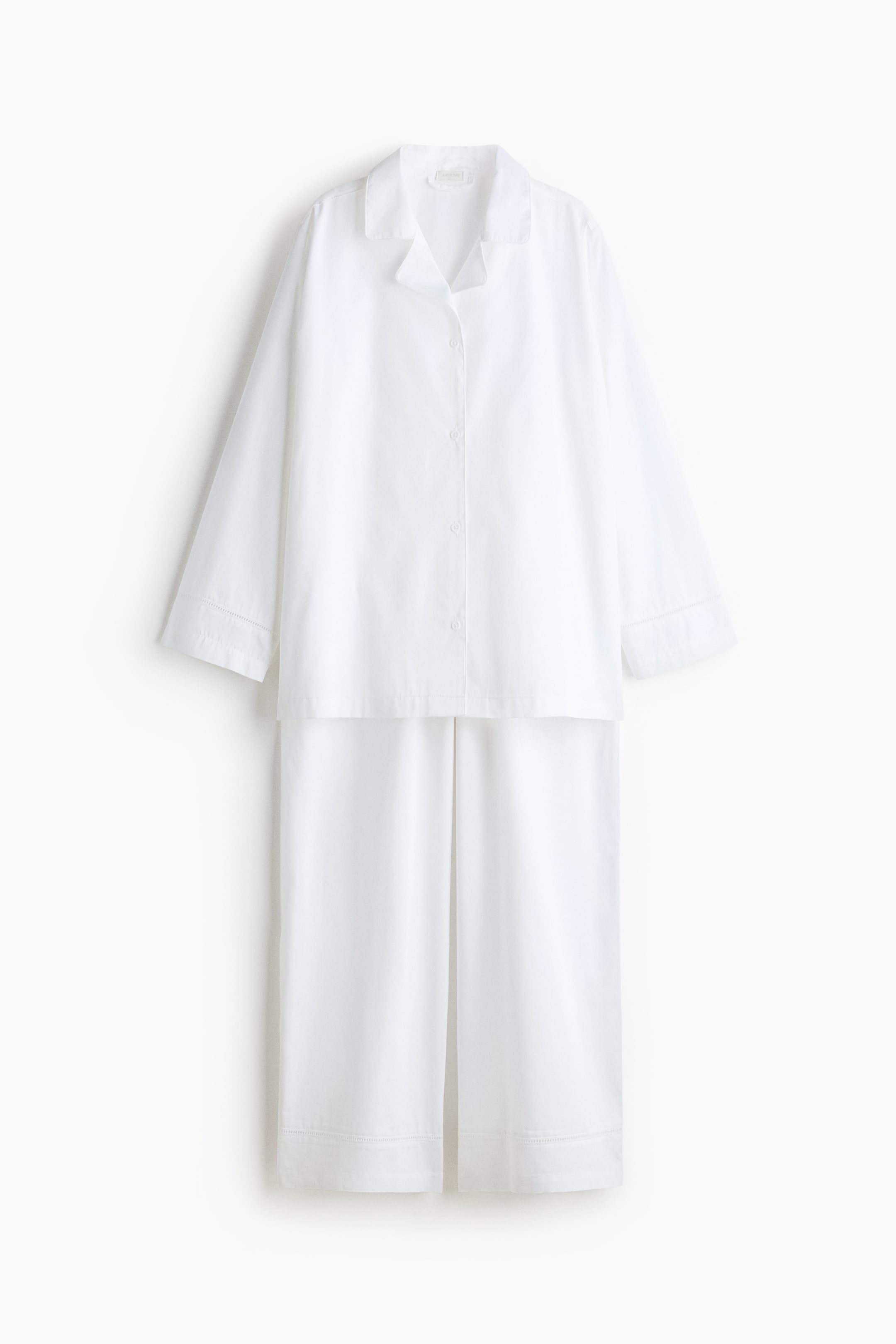 View larger image: A white pajama set is showcased in a front-facing view, comprising a long-sleeved button-down top with notch lapels and a button placket. Complementing the set are matching wide-leg trousers, adorned with delicate ladder-stitching details visible on the cuffs of the top and the hems of the trousers.