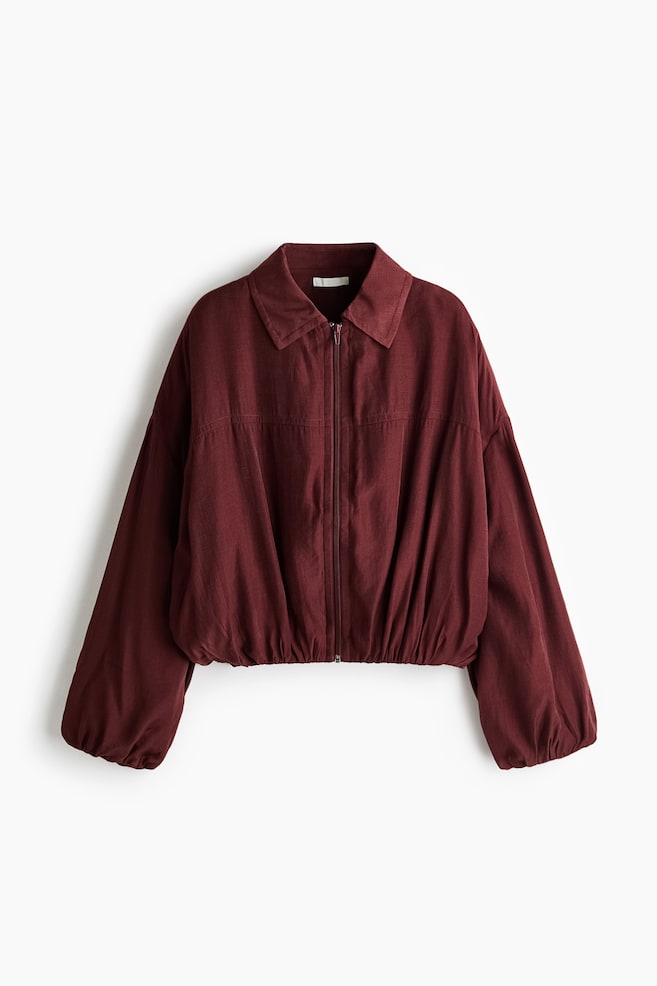 A front-facing burgundy jacket featuring a pointed collar, a full-length front zipper, and dropped shoulders with voluminous sleeves. The design includes a horizontal chest seam and an elasticized hem and cuffs, creating a gathered, blouson silhouette. - 1