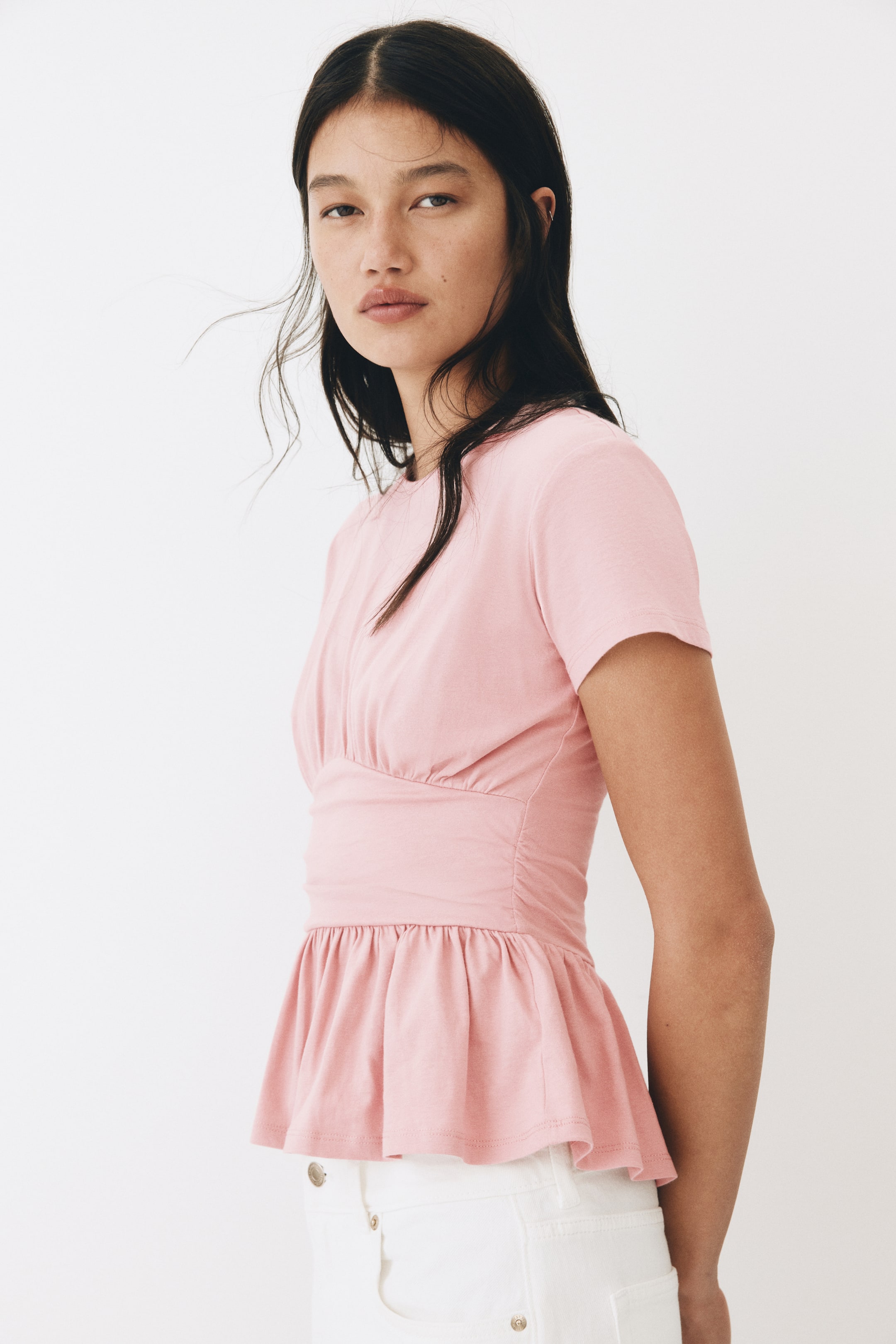 View larger image: A pink short-sleeved top featuring a round neckline, ruched detailing across the bust, and a fitted waist with a gathered peplum hem, viewed from a slight front-side angle.