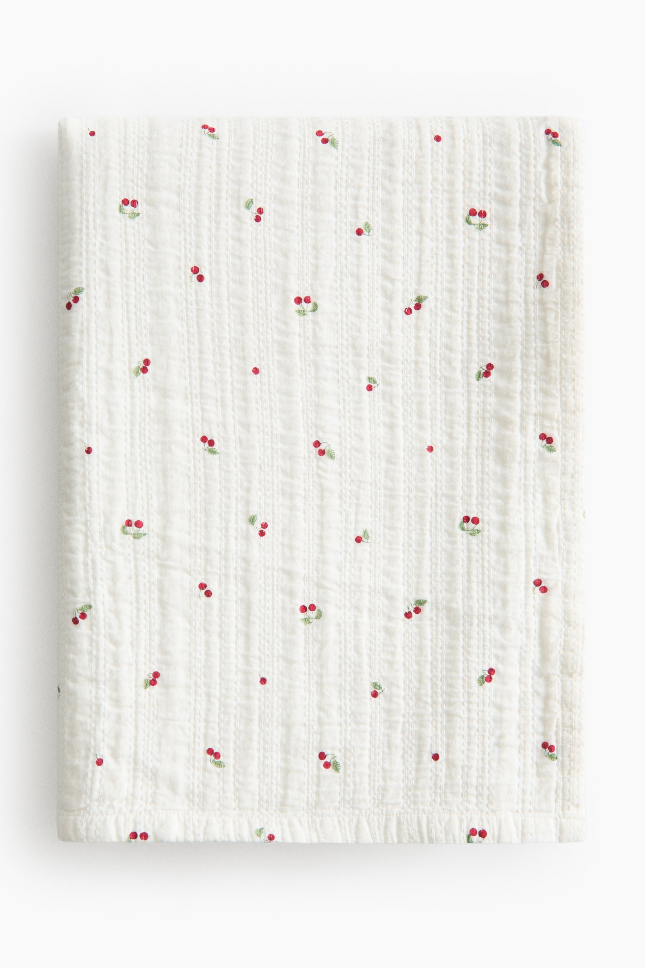 View larger image: A white quilted blanket showcases a scattered pattern of red cherries with green leaves. It features vertical stitched lines across its front surface.
