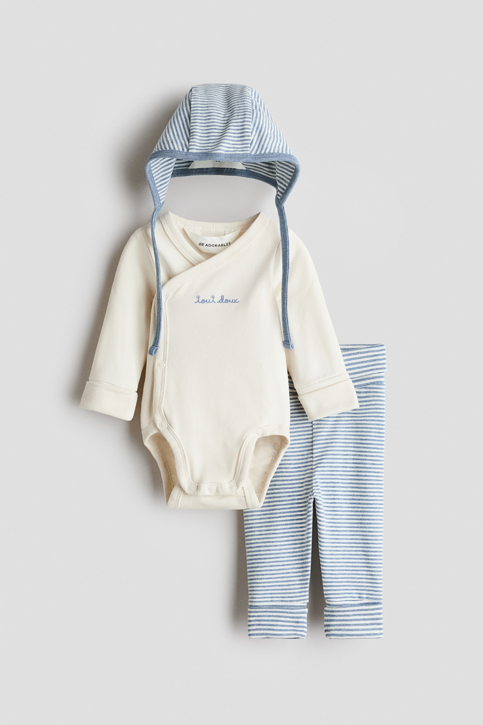 Un conjunto de ropa de bebÃ© de tres piezas presenta una vista frontal de un body cruzado de manga larga en color blanco roto, con bordado azul Â«tout douxÂ» en el pecho. El body se coordina con un pantalÃ³n de rayas horizontales azules y blancas y un gorro a juego de rayas con lazos para la barbilla.