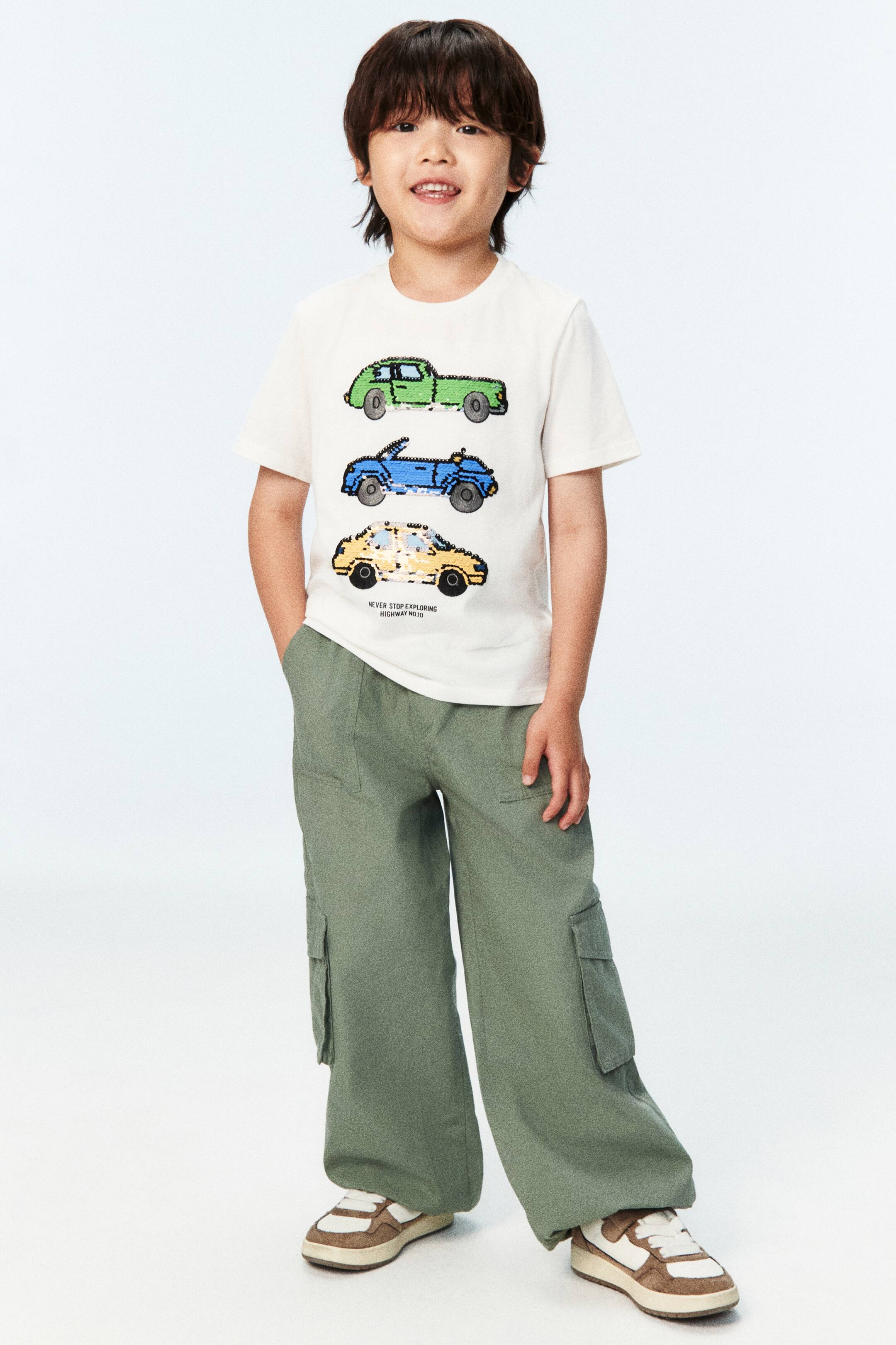 View larger image: The model is wearing an off-white short-sleeved t-shirt, adorned with three car motifs in green, blue, and yellow sequins on the front, accompanied by small text below. This t-shirt is styled with green wide-leg cargo pants and complemented by brown and white sneakers.