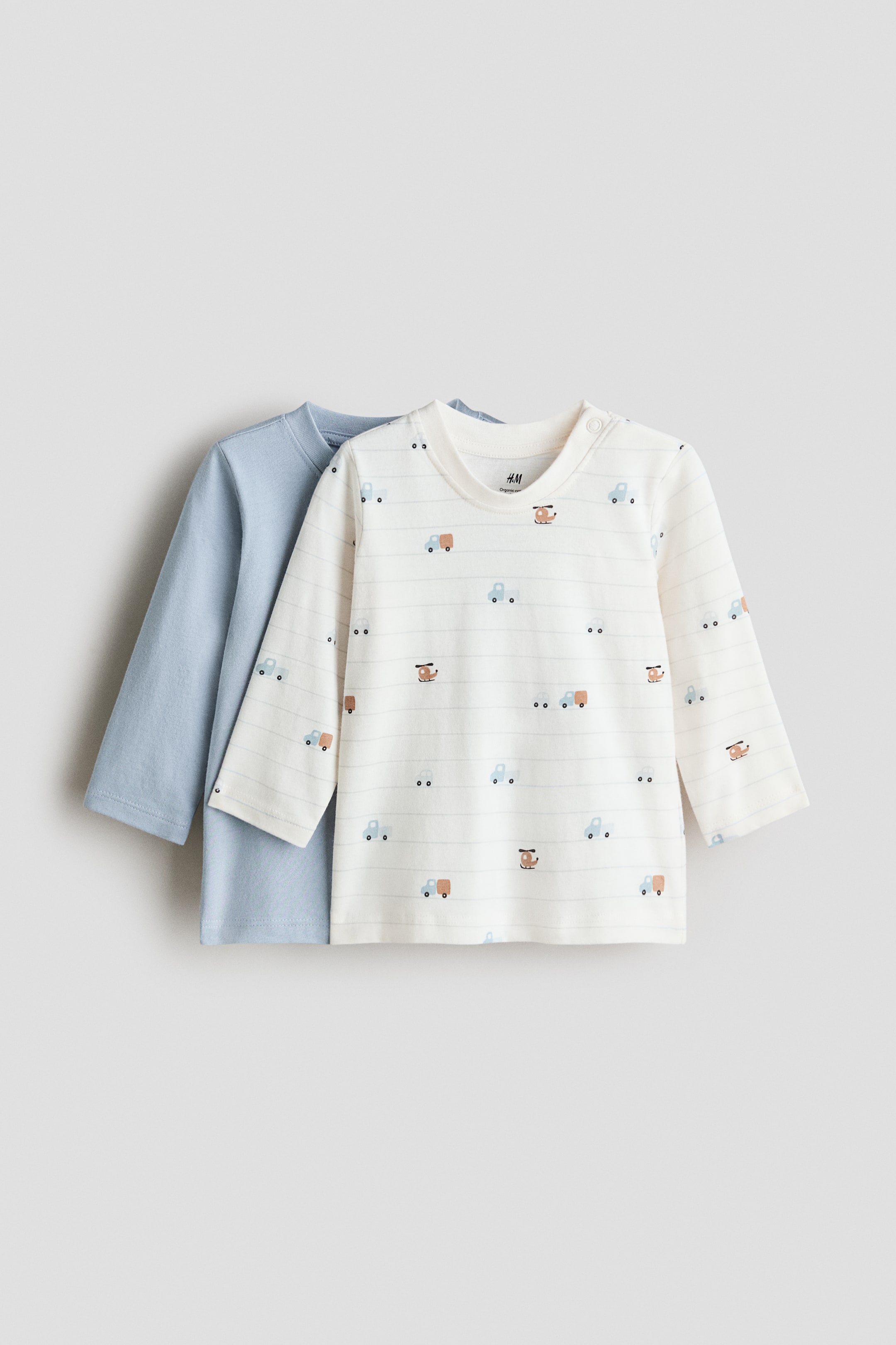 View larger image: A 2-pack of long-sleeved jersey tops shows two infant tops, both facing front. The top in the foreground is cream-colored, with horizontal light gray stripes and scattered vehicle motifs, and has a round neckline with two snap buttons on the left shoulder. The top positioned behind is solid light blue with a round neckline.