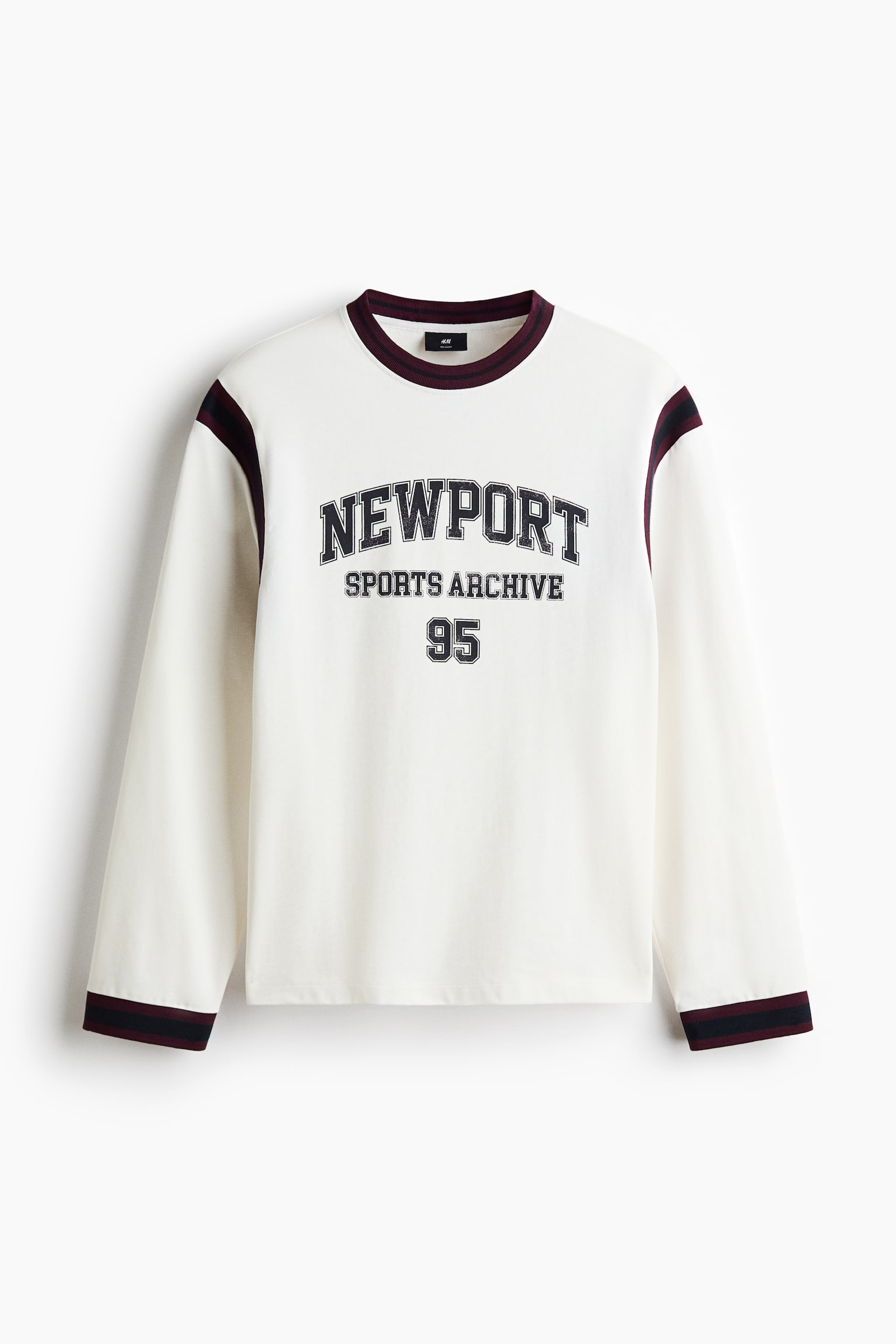 Un sweat-shirt Ã©cru Ã  manches longues est prÃ©sentÃ© de face, dotÃ© dâun col rond cÃ´telÃ© et de poignets assortis ornÃ©s de rayures bordeaux foncÃ© et bleu marine. La poitrine affiche un imprimÃ© graphique sombre de style varsity portant lâinscription Â« NEWPORT SPORTS ARCHIVE 95 Â» en lettres majuscules, tandis que des rayures sombres assorties soulignent les coutures dâÃ©paules.