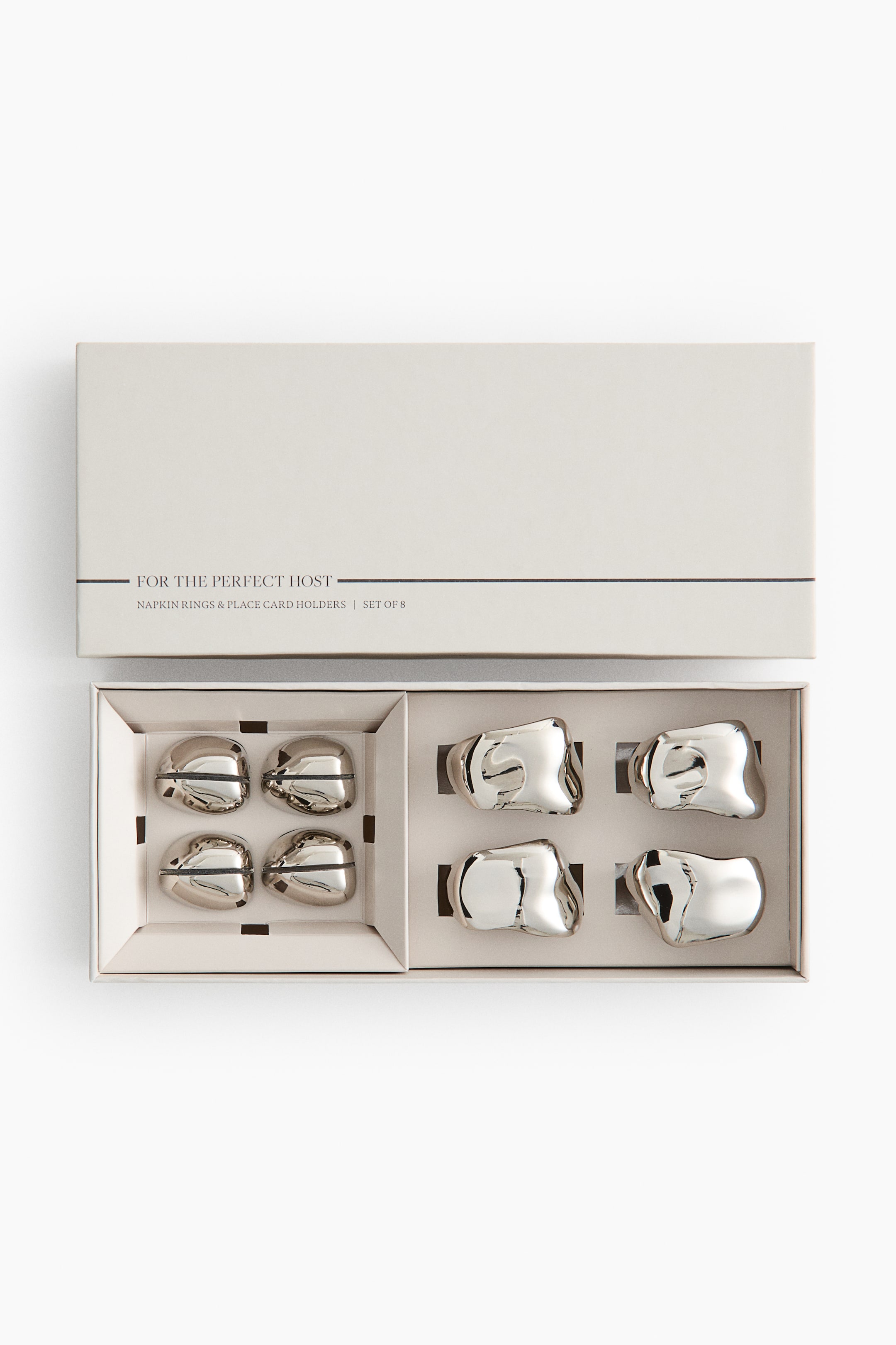 View larger image: A table setting set, made of polished stainless steel with an irregular design, is being presented in an open box. Four napkin rings are occupying the right compartments, while four smaller place card holders, featuring a top slot, are occupying the left compartments.