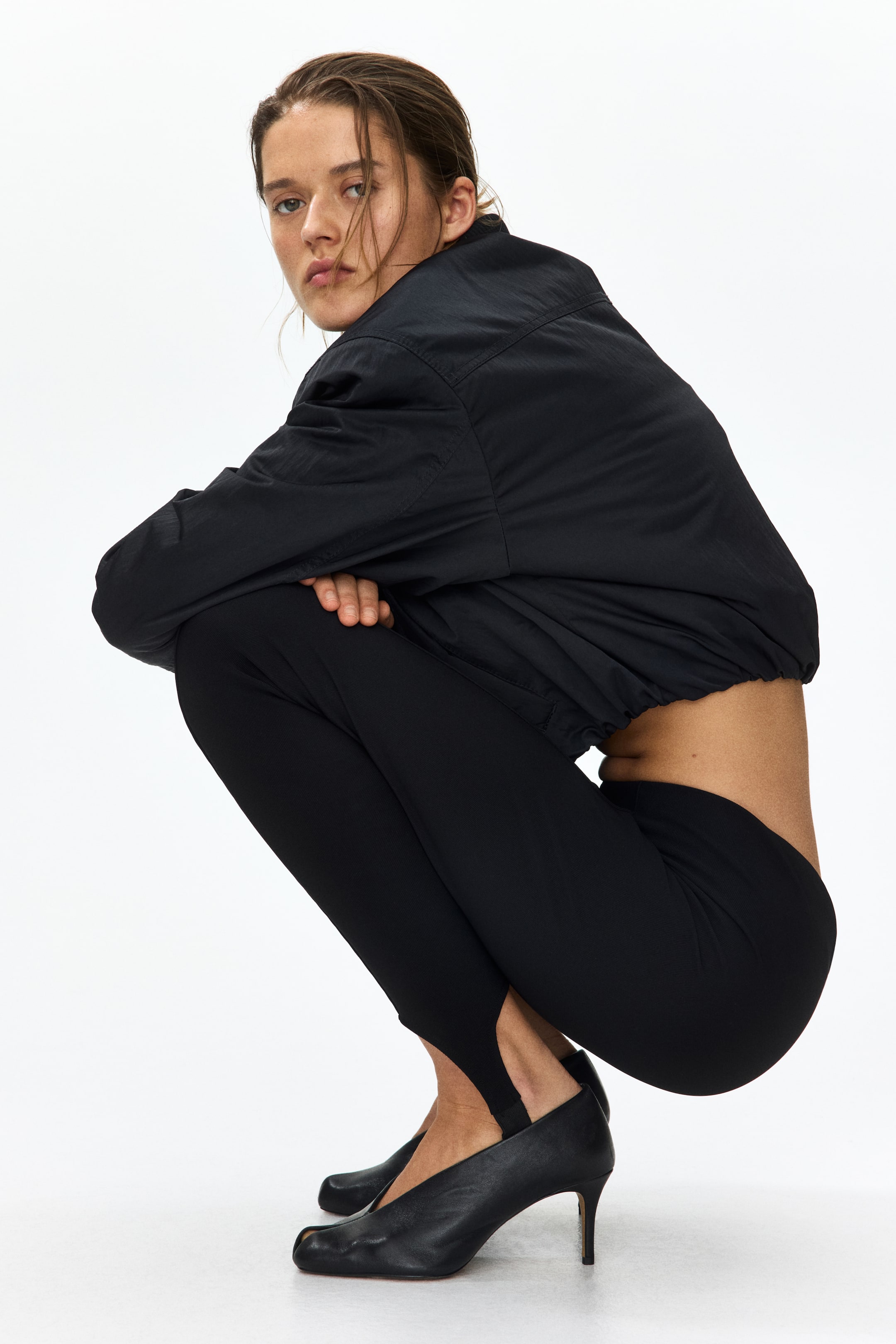 View larger image: A black bomber jacket features a stand collar, elasticated cuffs, and a gathered elasticated hem. Black form-fitting stirrup leggings stretch over the legs, with the stirrups hooking under the heels. Black leather split-toe heels are visible from a side-front angle, featuring a stiletto heel.