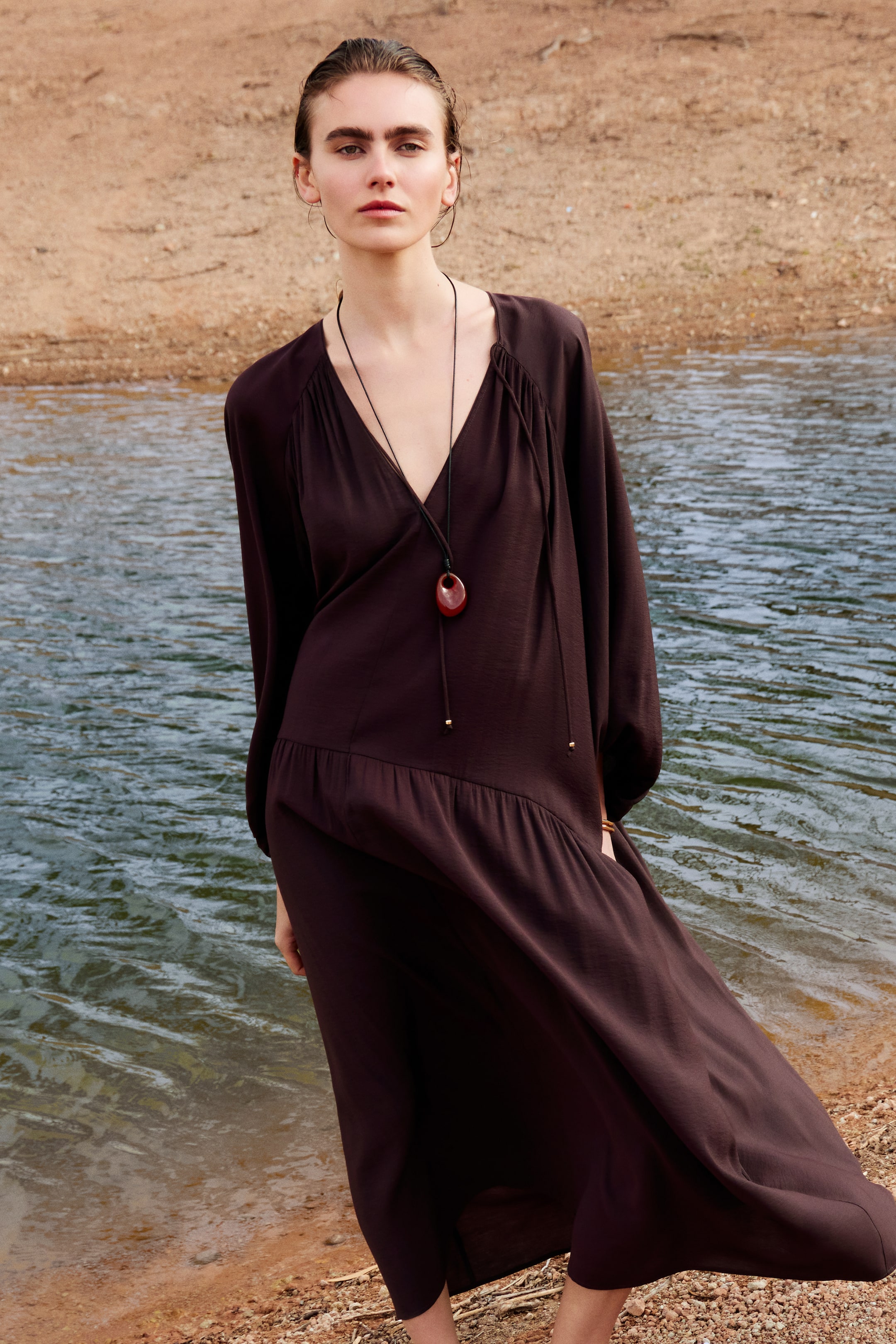 View larger image: The model is wearing an oversized dark brown dress, featuring a V-neckline with spaghetti ties at the top, long voluminous sleeves with gathers at the shoulders, and a curved gathered seam over the hips. A necklace with a reddish-brown pendant rests on the dress.