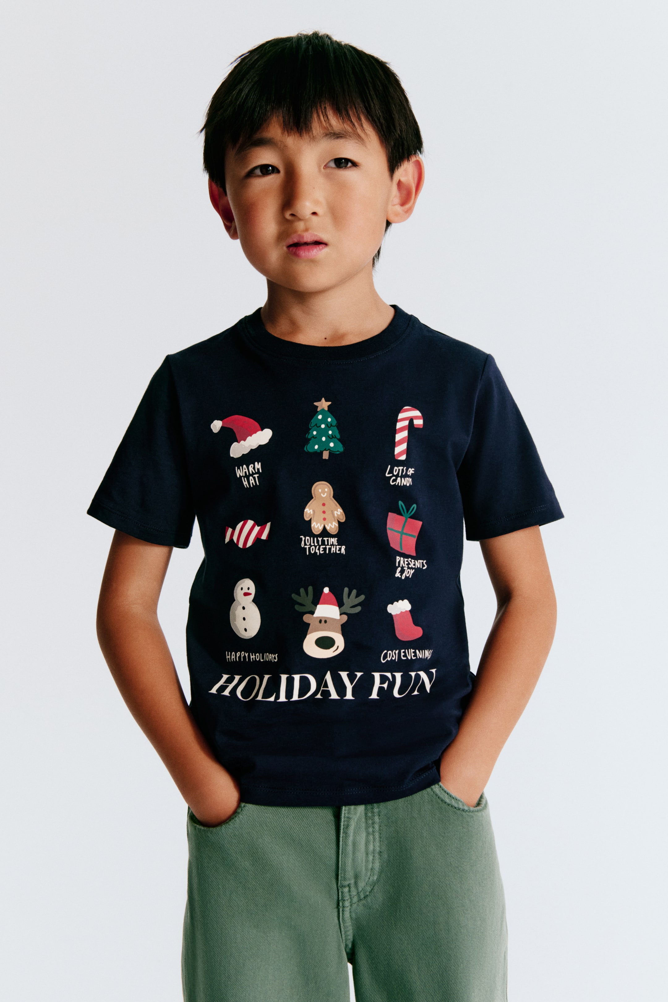View larger image: A dark navy blue cotton jersey T-shirt is shown from the front, featuring a round, rib-trimmed neckline and short sleeves with gently dropped shoulders. The chest displays a printed design of various holiday-themed illustrations and text, culminating in the words "HOLIDAY FUN" in large white lettering at the bottom.