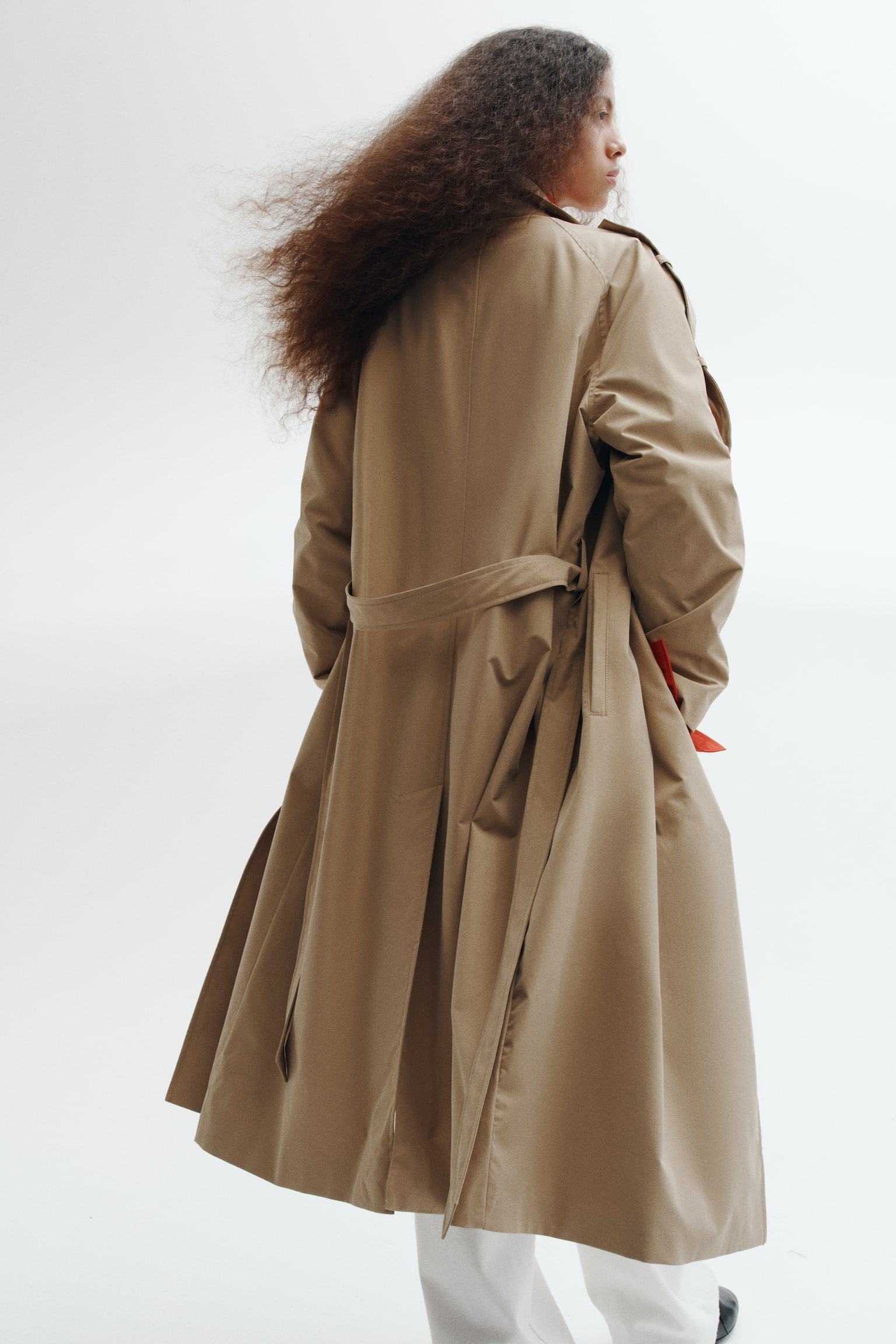 A tan double-breasted trench coat features notch lapels, dark brown buttons, shoulder tabs, and adjustable sleeve straps. A matching belt is casually tied around the waist, with two side pockets in view. - 1