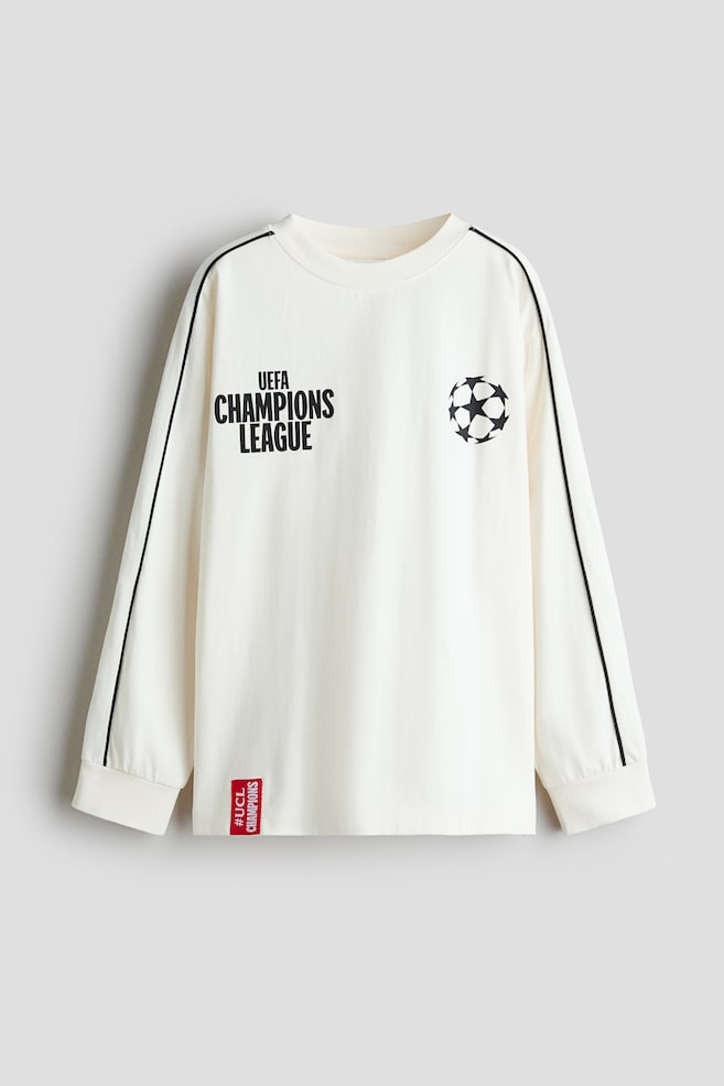 Un t-shirt Ã  manches longues blanc cassÃ© est prÃ©sentÃ© de face, avec un col rond cÃ´telÃ© et des poignets ornÃ©s dâun fin passepoil noir le long des manches. Le haut affiche le texte noir Â« UEFA CHAMPIONS LEAGUE Â» sur la poitrine droite, un logo starball noir sur la poitrine gauche, ainsi quâune petite Ã©tiquette tissÃ©e rouge prÃ¨s de lâourlet infÃ©rieur. - 1